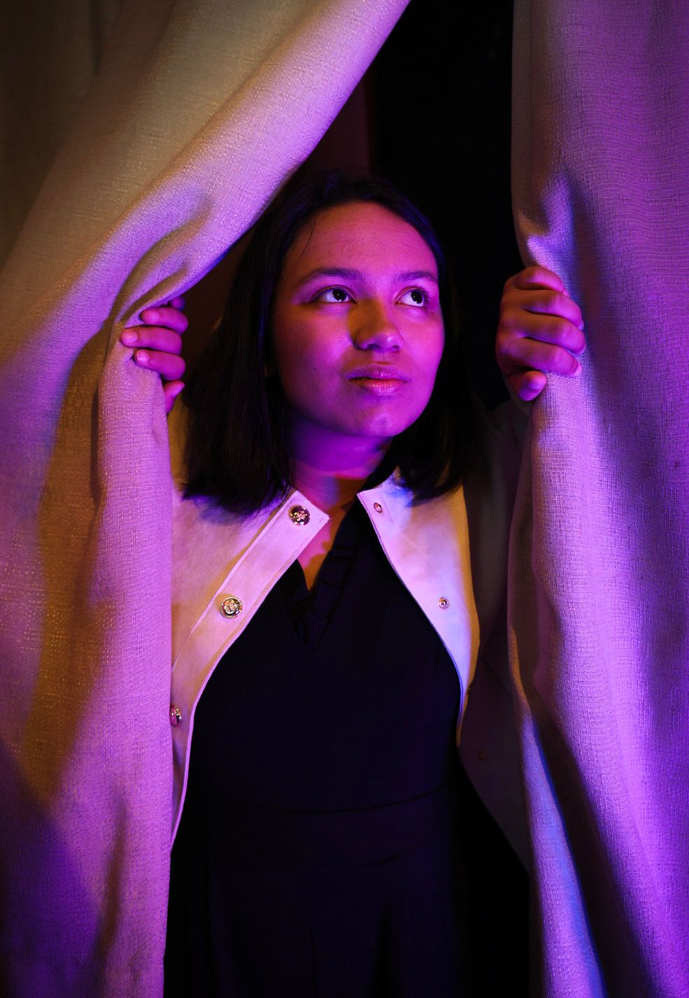 Senior portrait of a girl peeking out from behind a curtain at The Roxie Theatre, there are blue and purple lights cascading across.