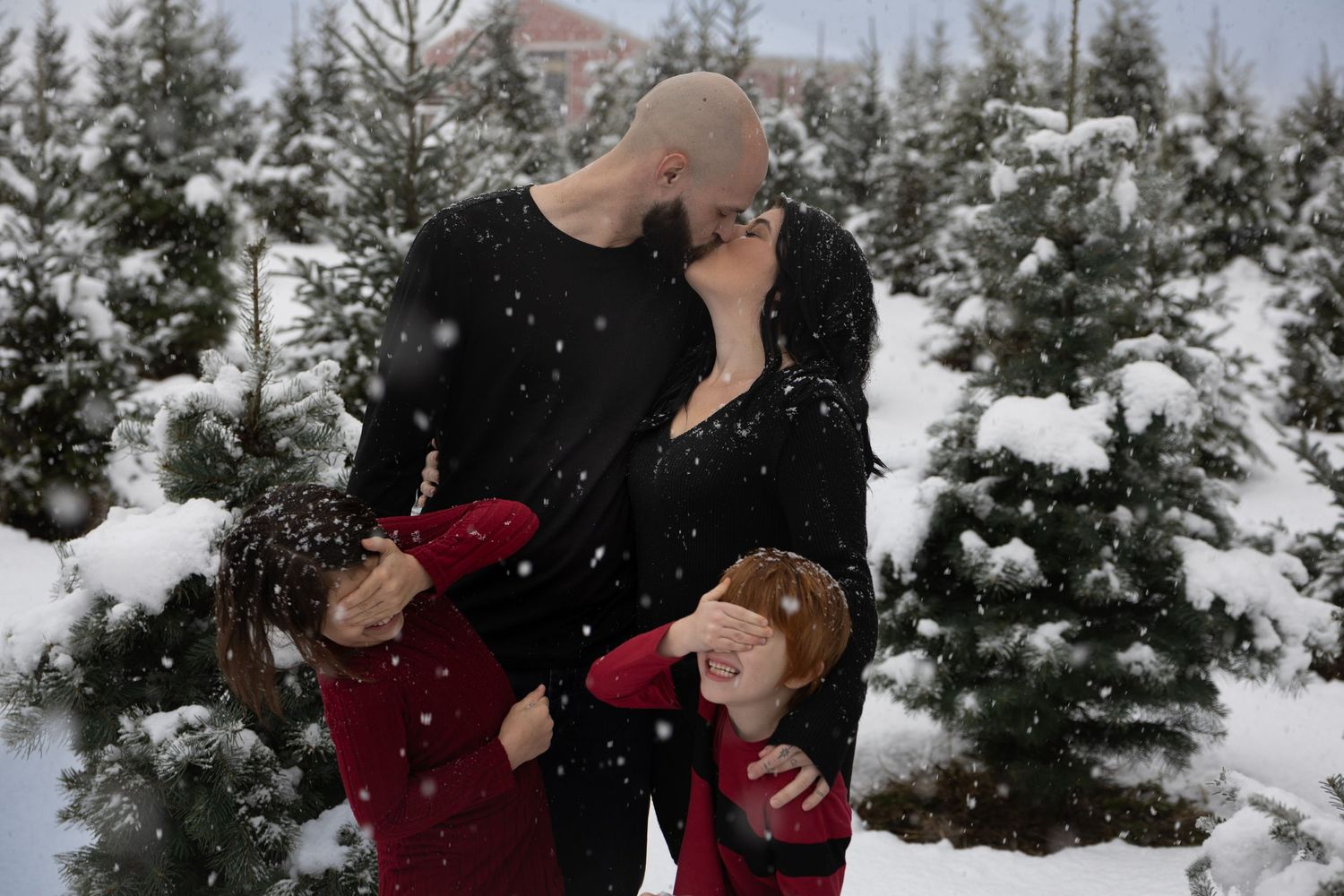 Family of 4 at a snowy Copenhaver Tree Farm in Missoula, Montana. Adults are kissing while children cover their eyes and make silly faces.