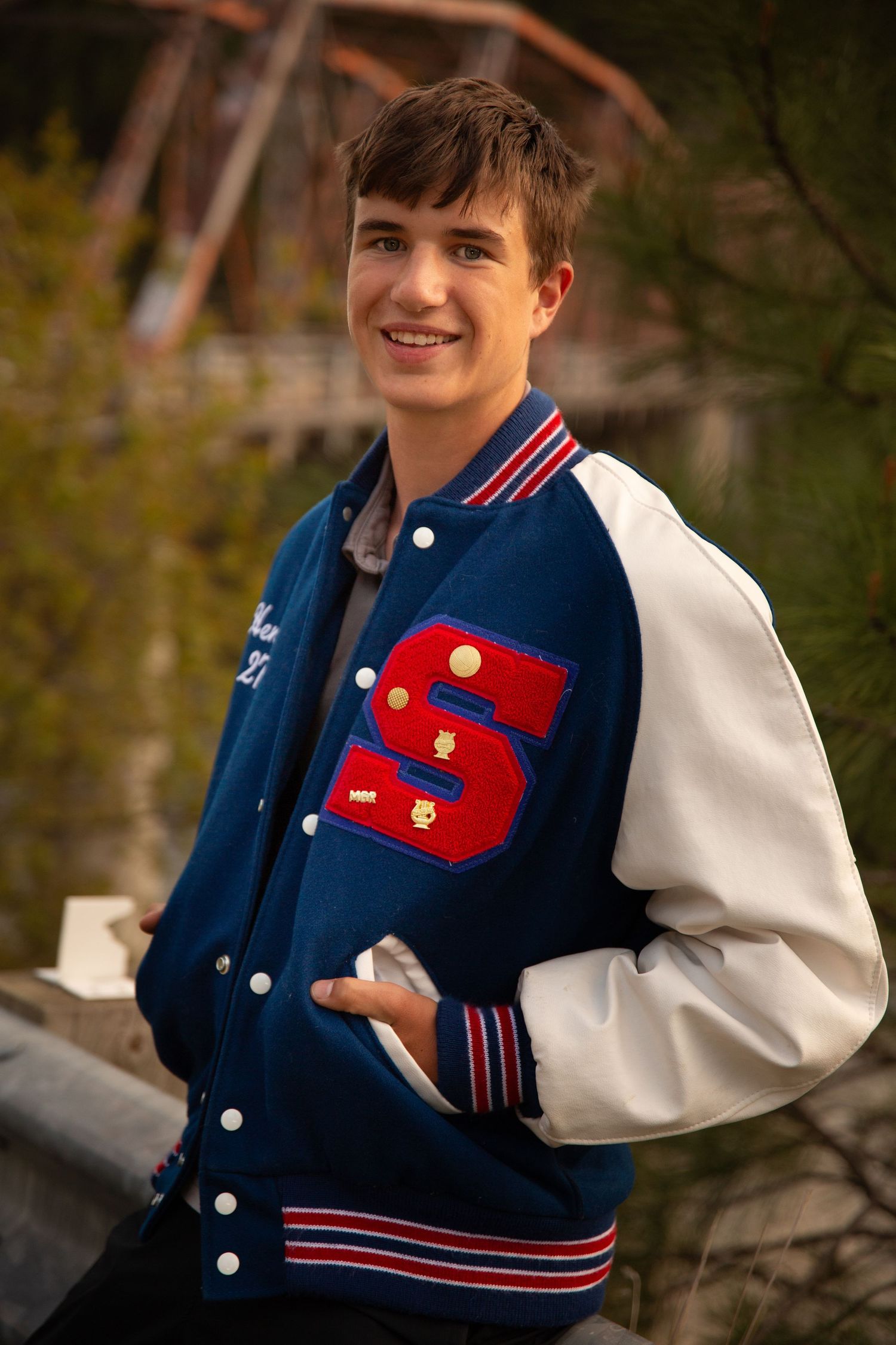 Senior portrait of young man wearing his high school letterman jacket during sunset, and there is the Lozeau, Mt bridge in the background.