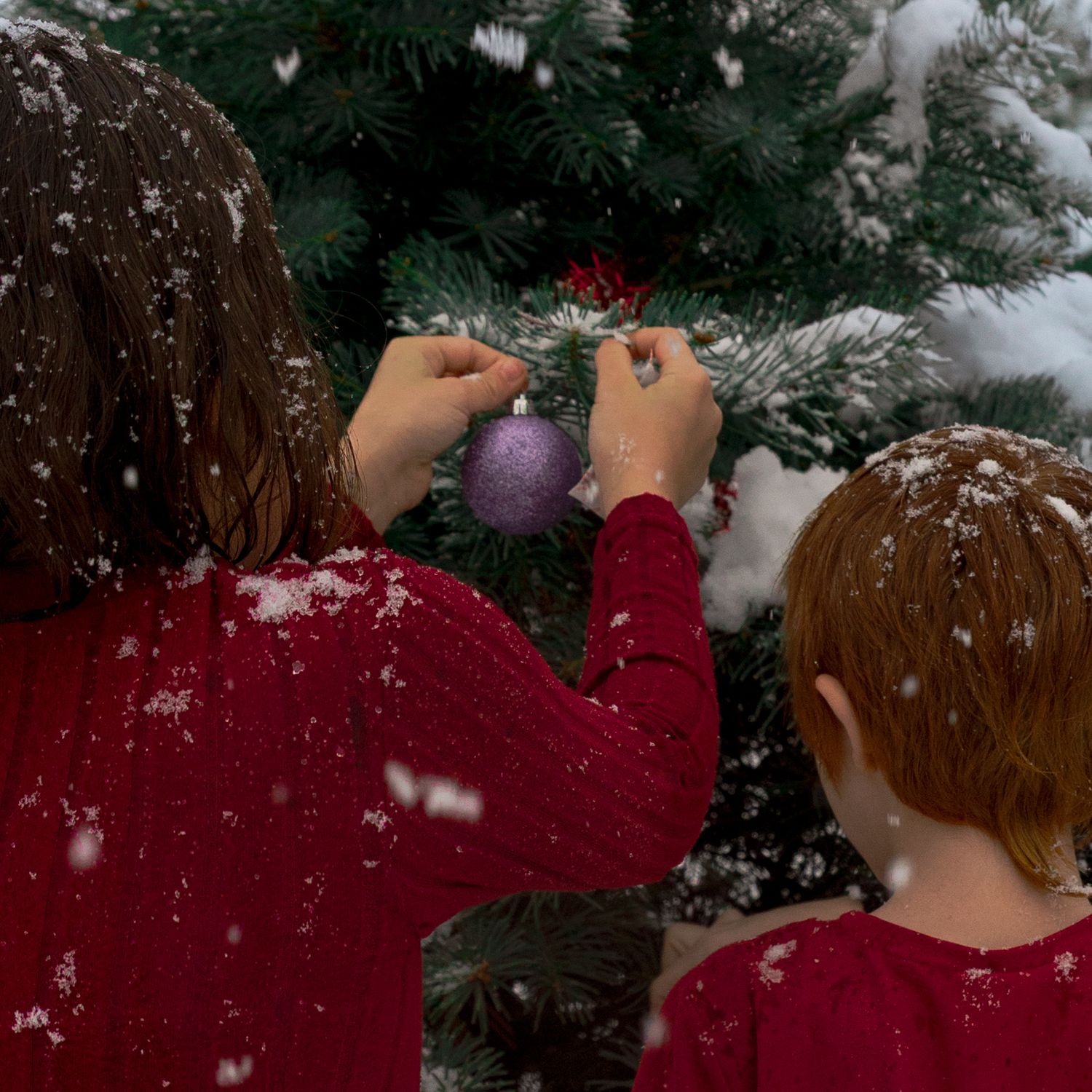 Two children, 1 boy and 1 girl, hanging up a Christmas ornament on a snow covered Christmas tree.