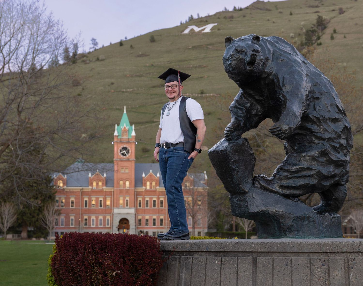Graduate of the University of Montana standing next to a statue of the grizzly bear mascot University Hall and Mount Sentinel in the back.