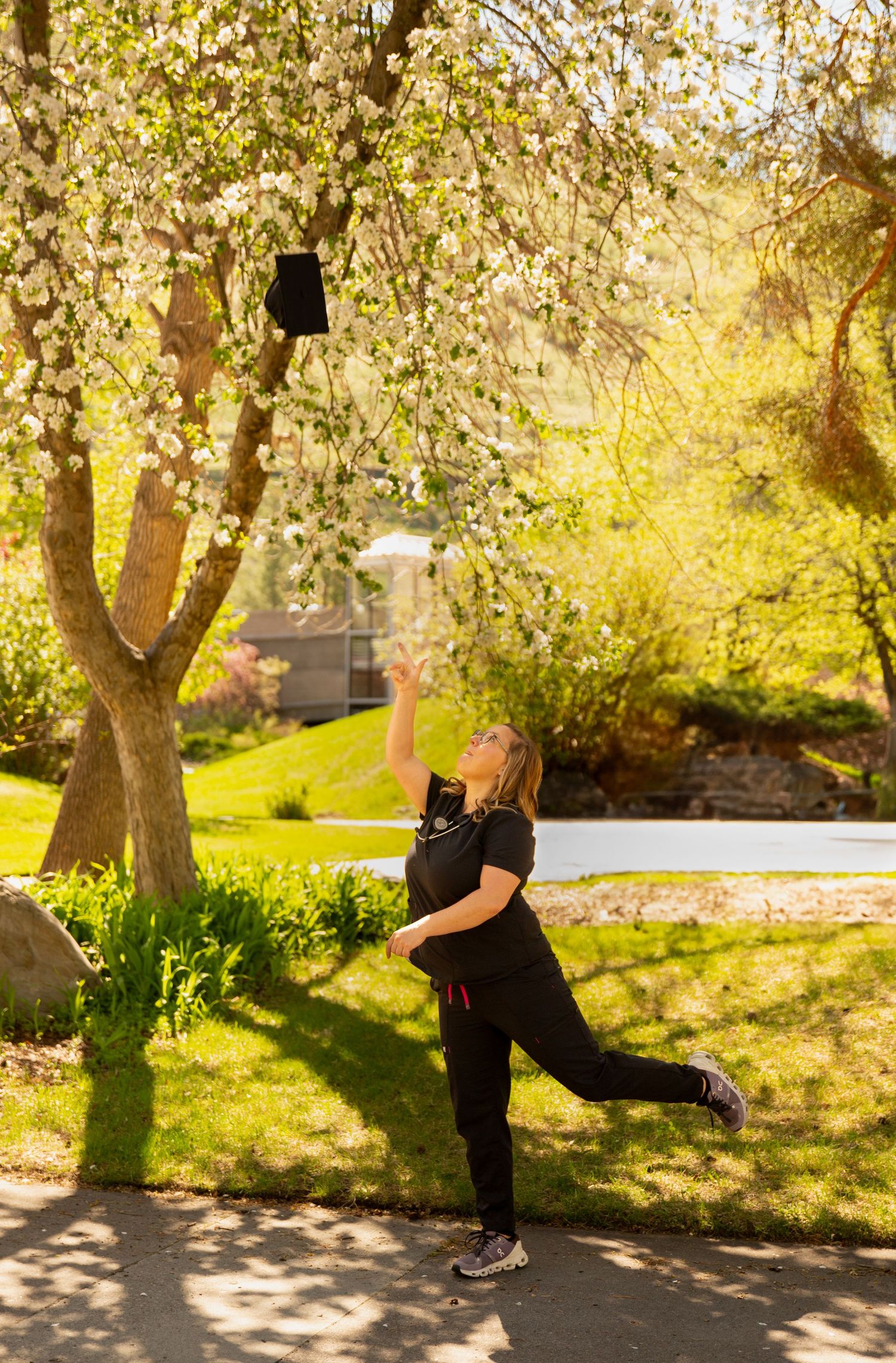 University of Montana nursing graduate tossing her cap high into the air. She is dressed in black scrubs, with a blooming tree in the back.