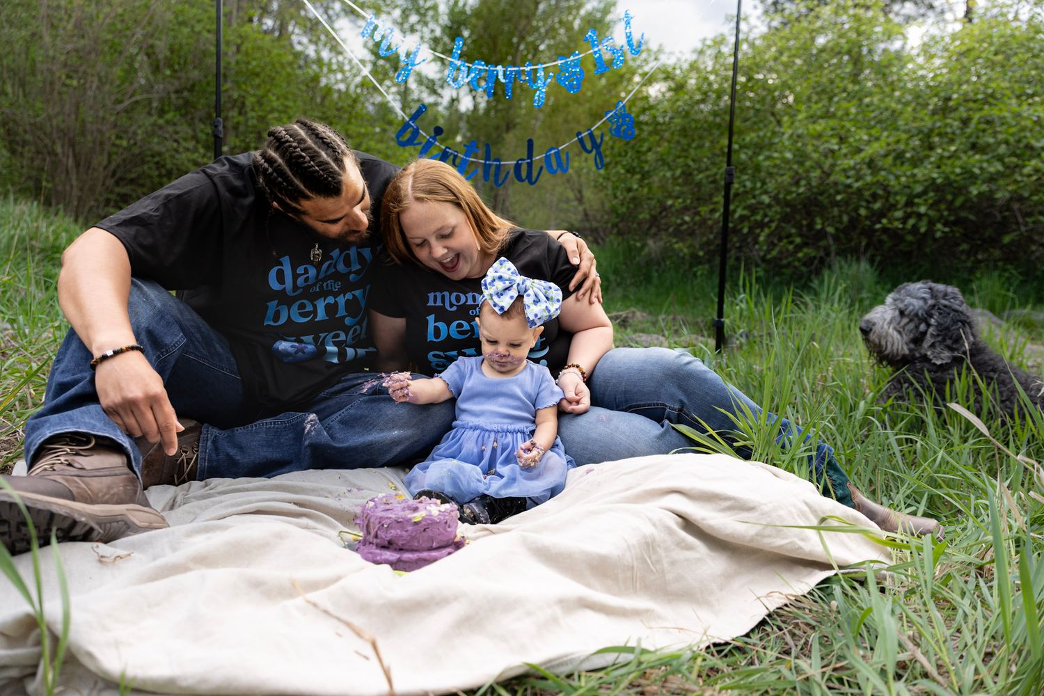 Family portrait of 3, toddler turning 1 year old, family surrounds berry cake, banner reads Berry 1st Year grey/black dog in the background.
