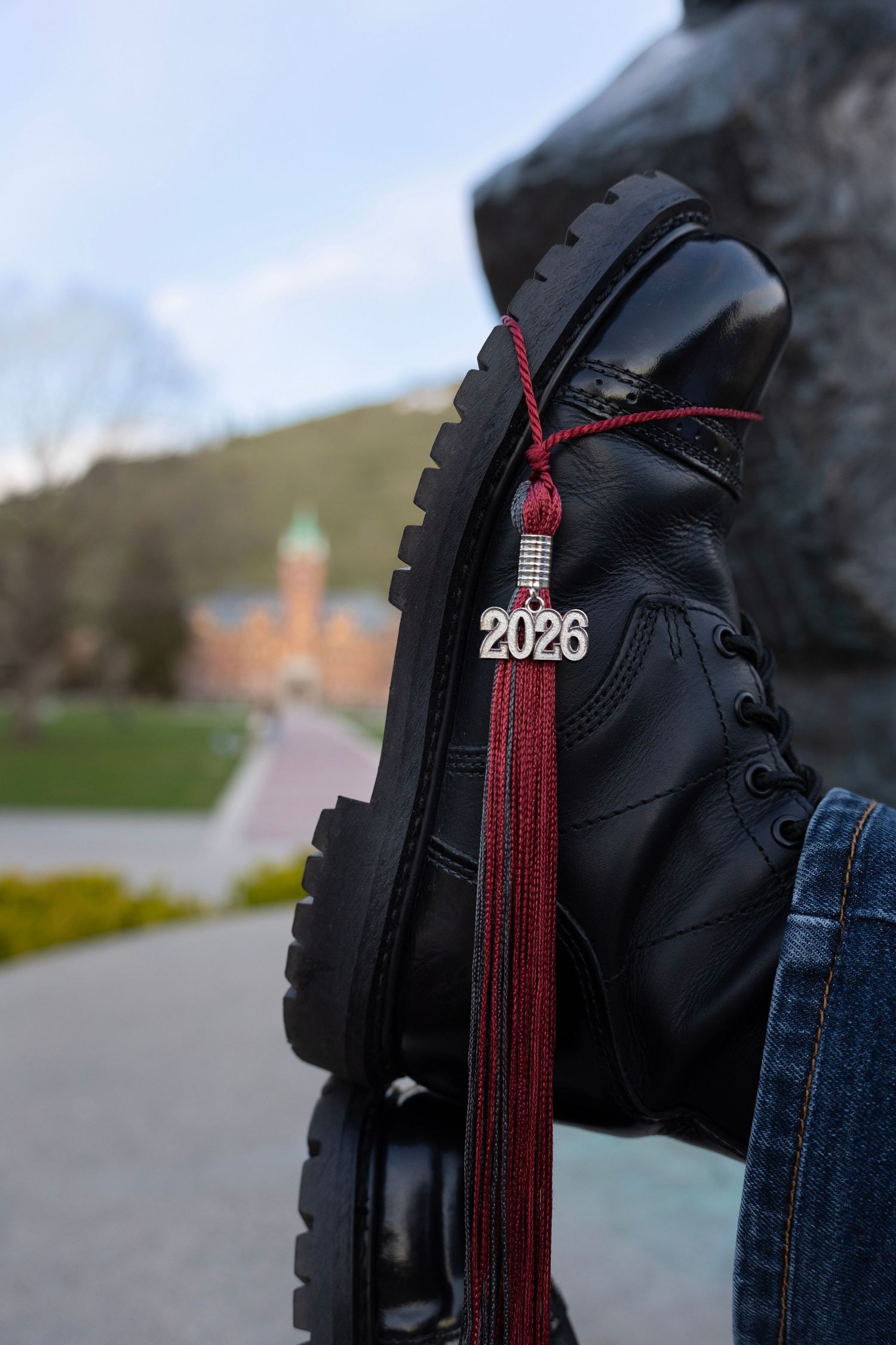 Black leather boots with a graduation tassel hanging from the toe and the University of Montana Dean's Hall in the background.