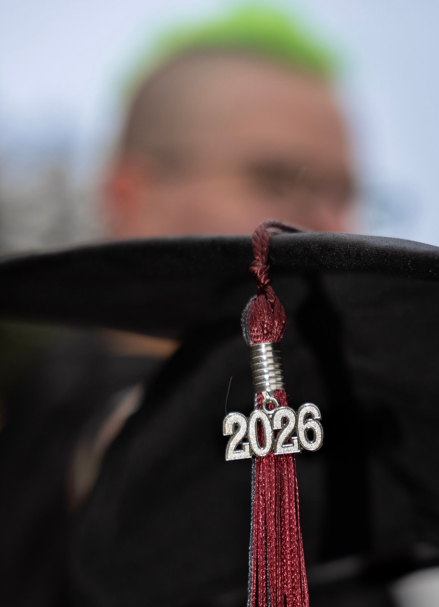 Graduate with green hair is holding cap and tassel very close to the camera to feature the 2026 metal piece on the tassel.