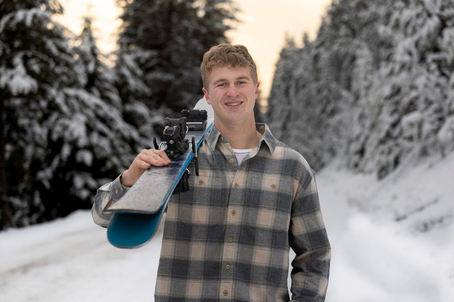Senior portrait of young man holding skis with a snowy background in Montana and the sun peaking up behind him.