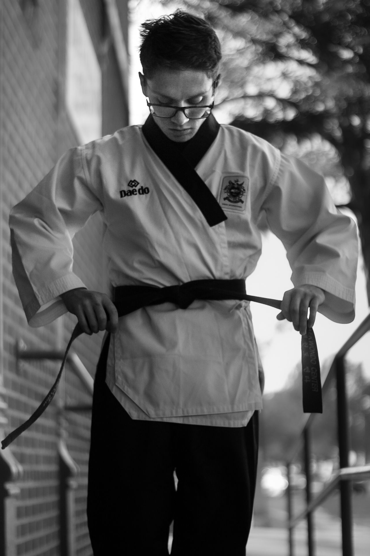 Senior portrait of young man wearing taekwondo dobok and tying his belt at his waist. Photo is in black/white, daytime, downtown Missoula.