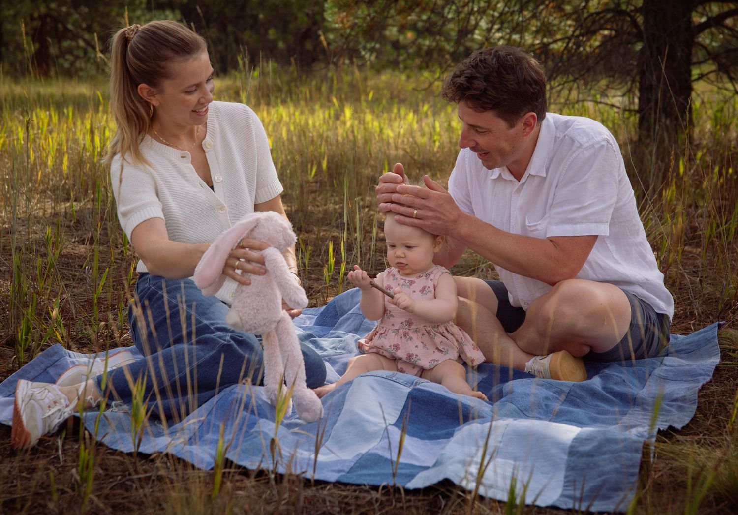 Portrait, parents and baby sitting on a blue patchwork blanket in a field. Mom is holding stuffed rabbit, while dad plays with baby's hair.