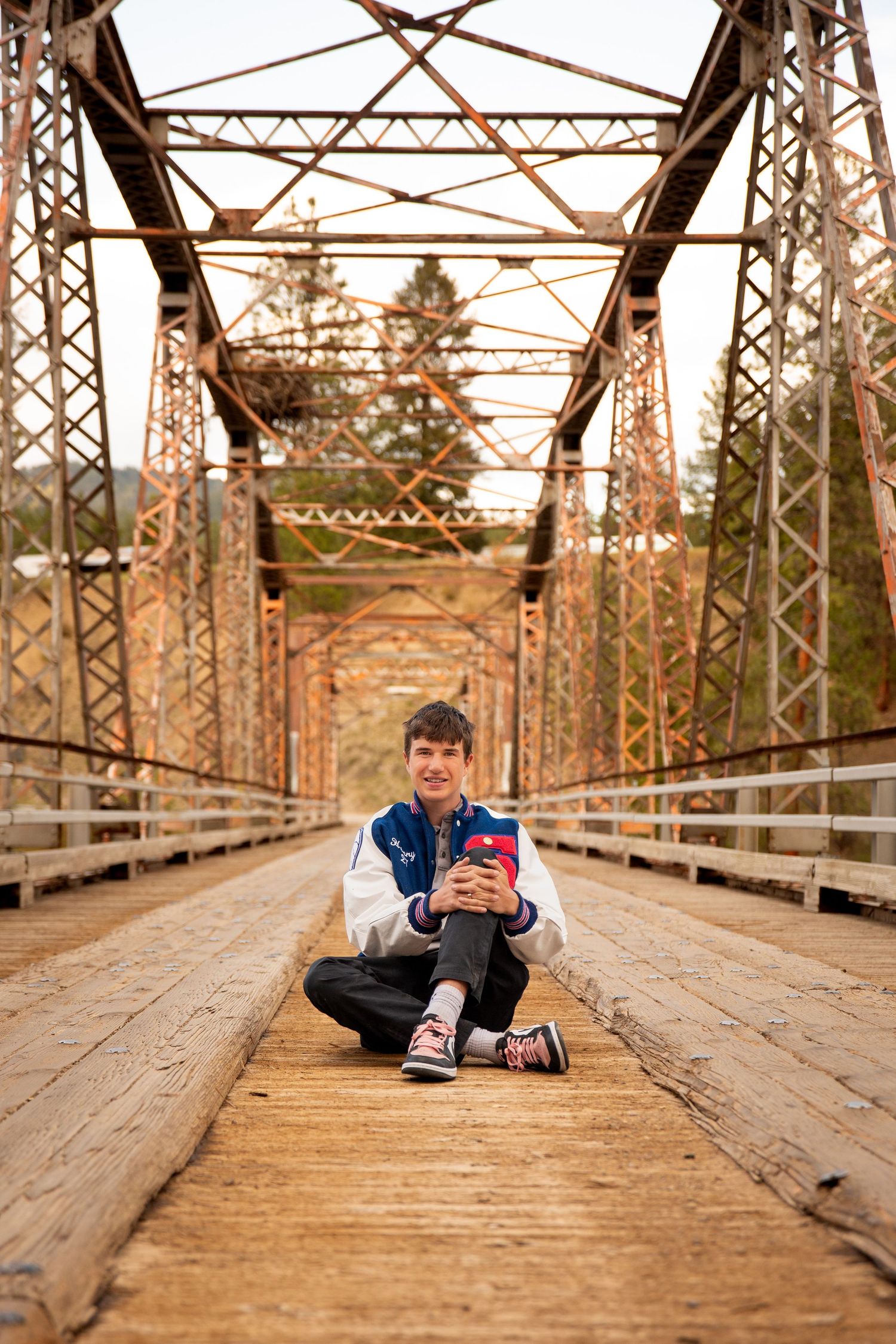 Senior portrait of young man sitting in the middle of the Lozeau bridge while wearing his letterman jacket on a sunny day.