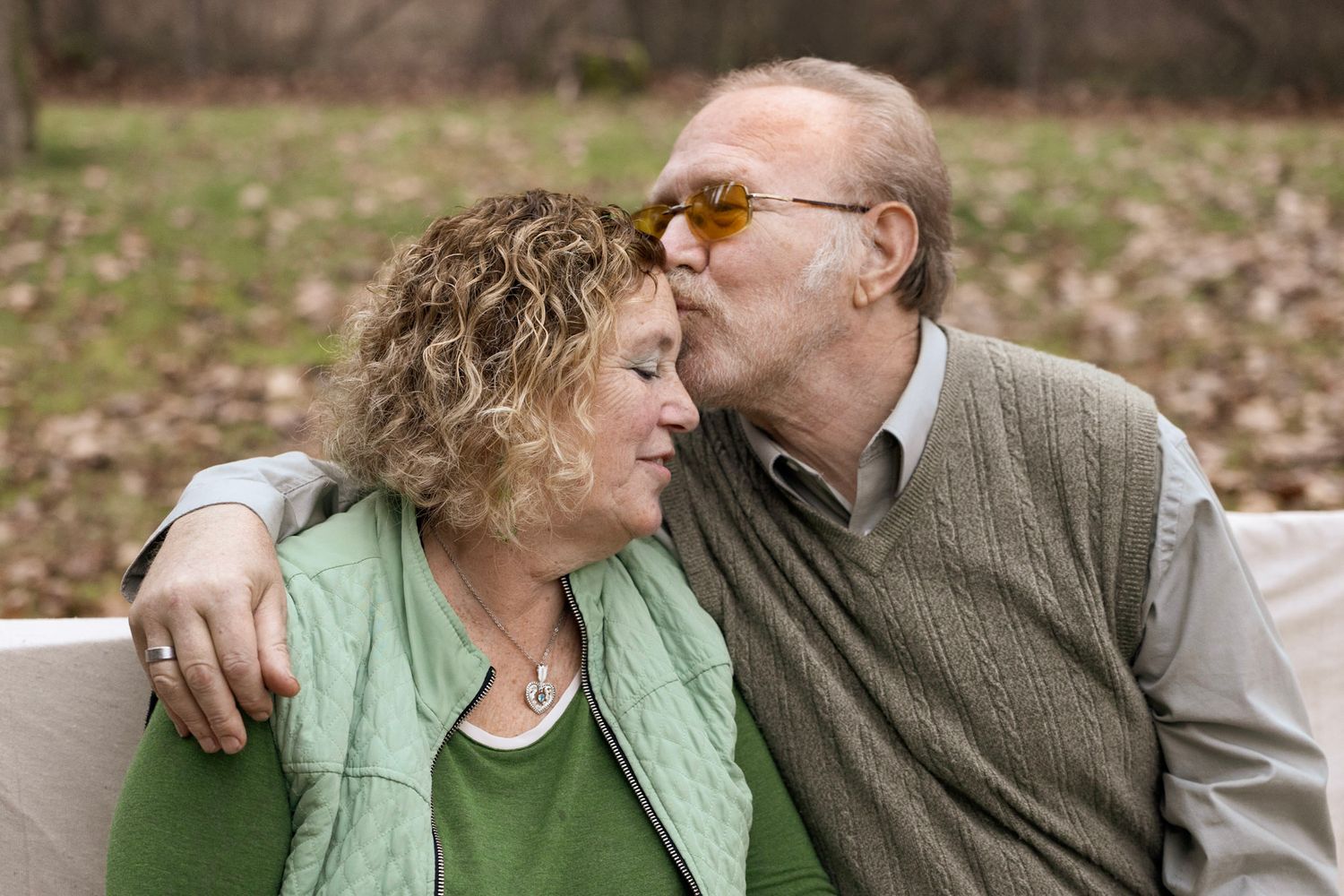Portrait of 2 grandparents, man is kissing wife's forehead while seated on a bench outside, located in Superior, Montana.
