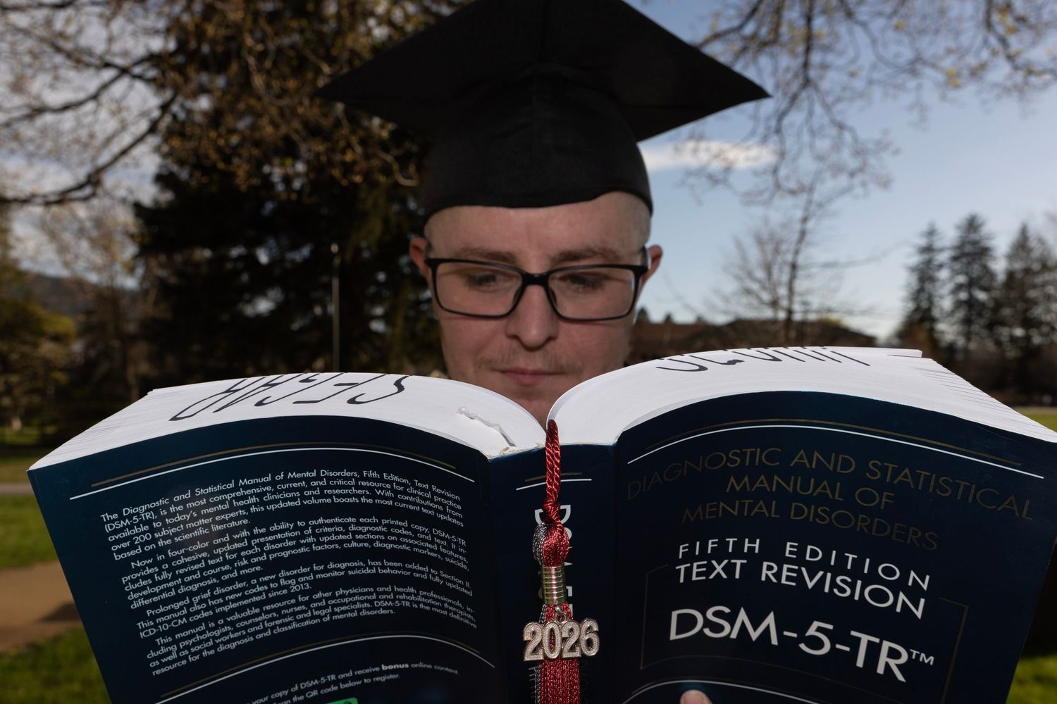 University of Montana graduate wearing black cap. Holding DSM-5 and the bookmark in the middle of the pages is the 2026 maroon tassel.