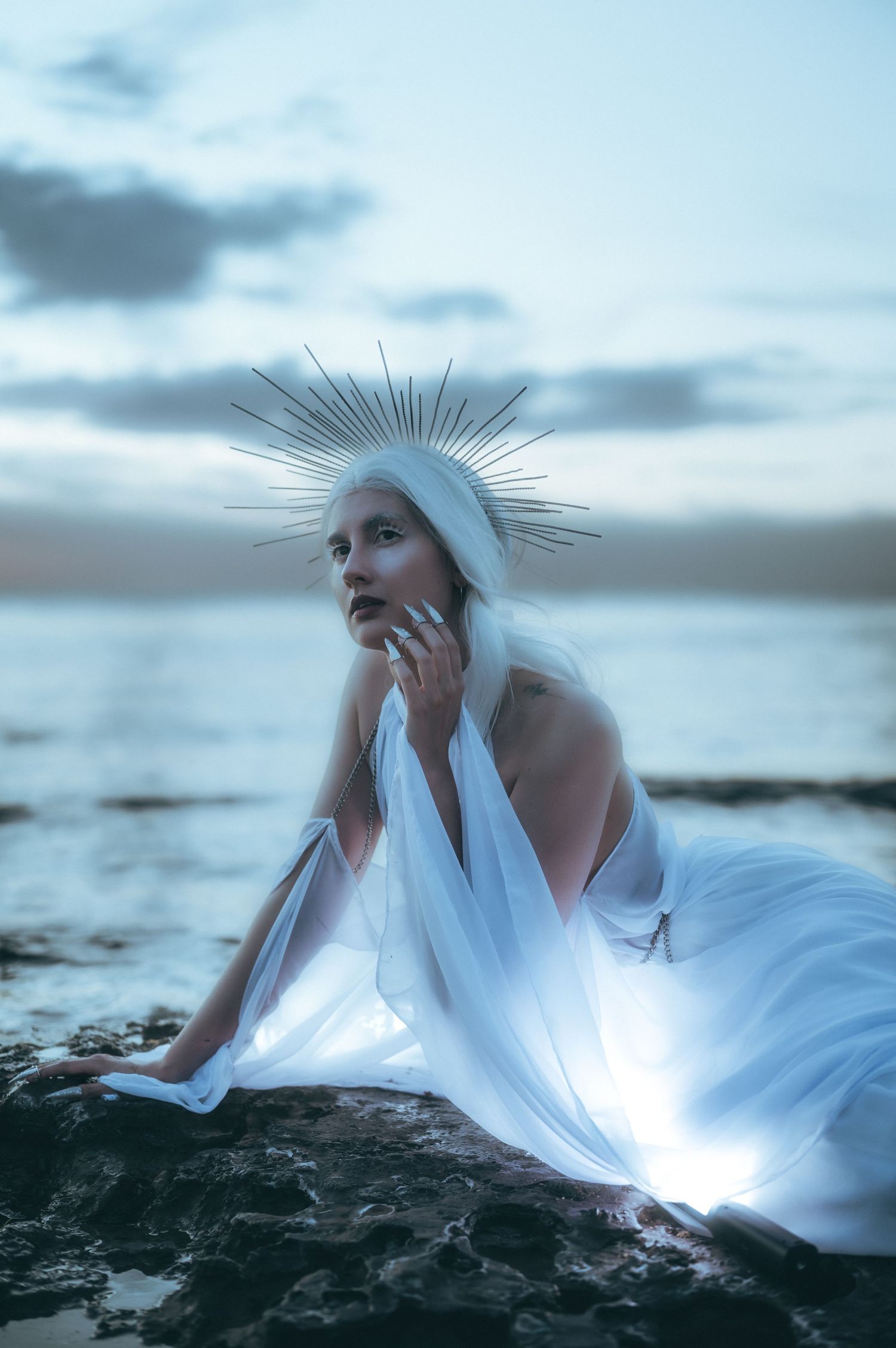 Artistic portrait featuring white fabric and crown-like headpiece against dramatic coastal backdrop.