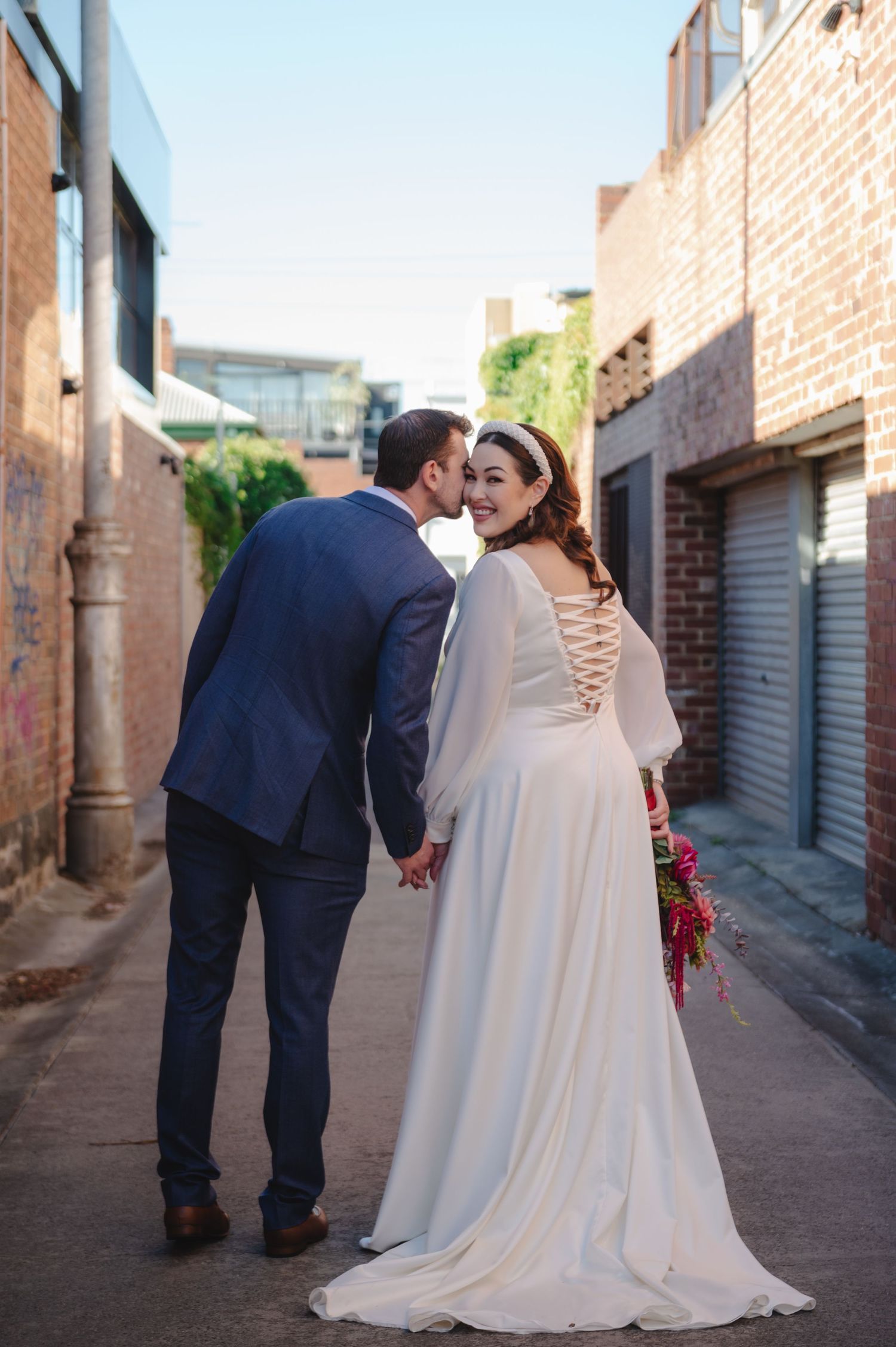 A romantic wedding couple sharing an intimate moment in an urban alleyway with brick walls and sunlight.