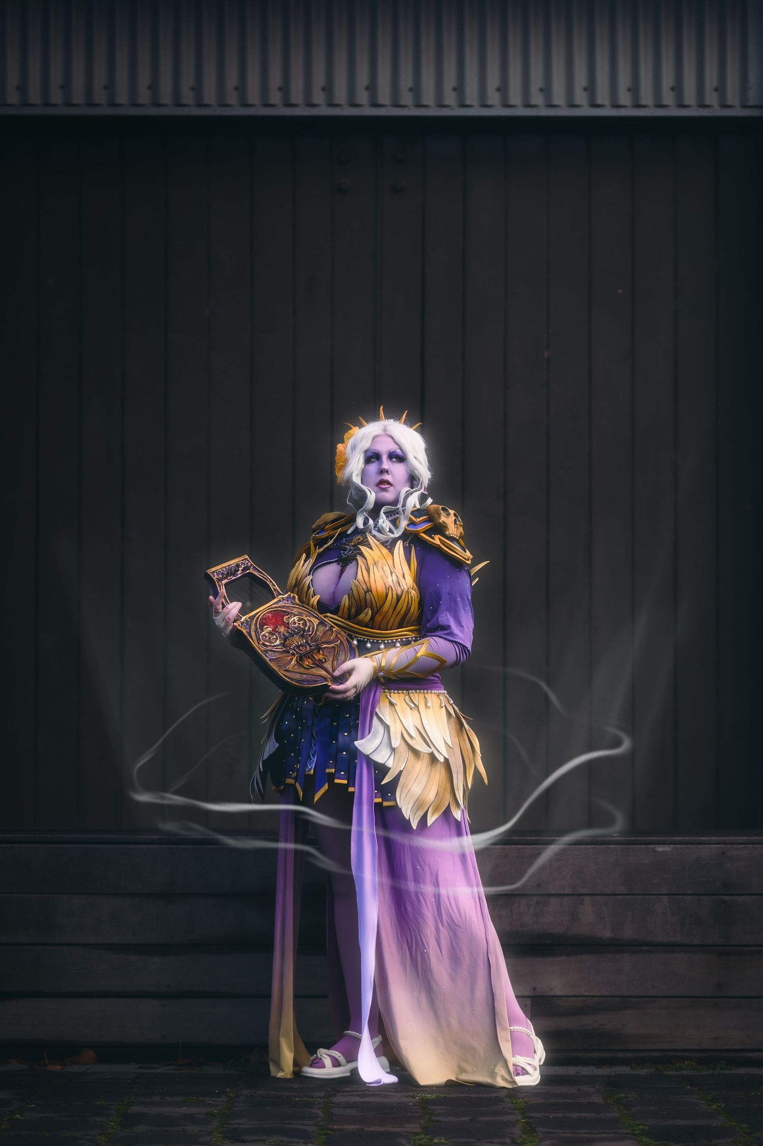 Cosplayer in flowing purple dress poses dramatically against dark background with fantasy props.