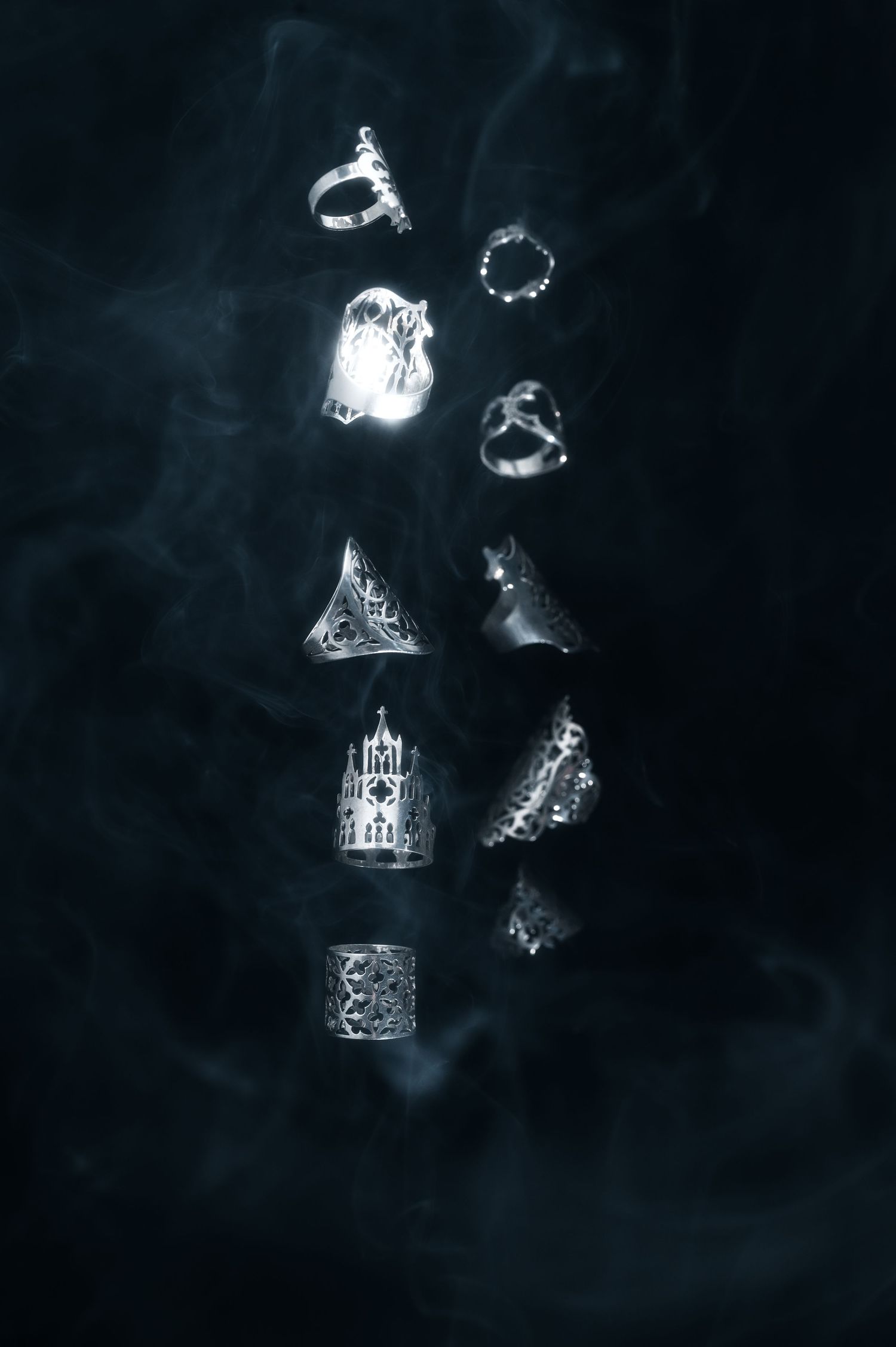 Collection of glowing silver rings and jewelry pieces floating against a smoky dark background.