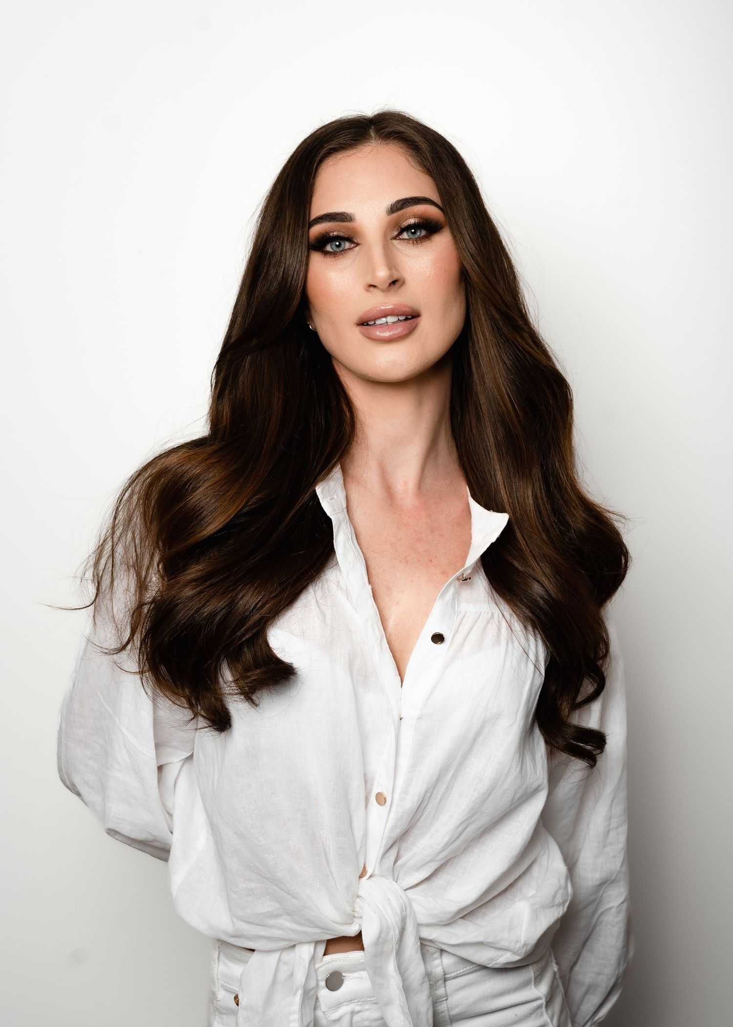 Young woman with long dark wavy hair wearing a white linen shirt, posing against a white background.
