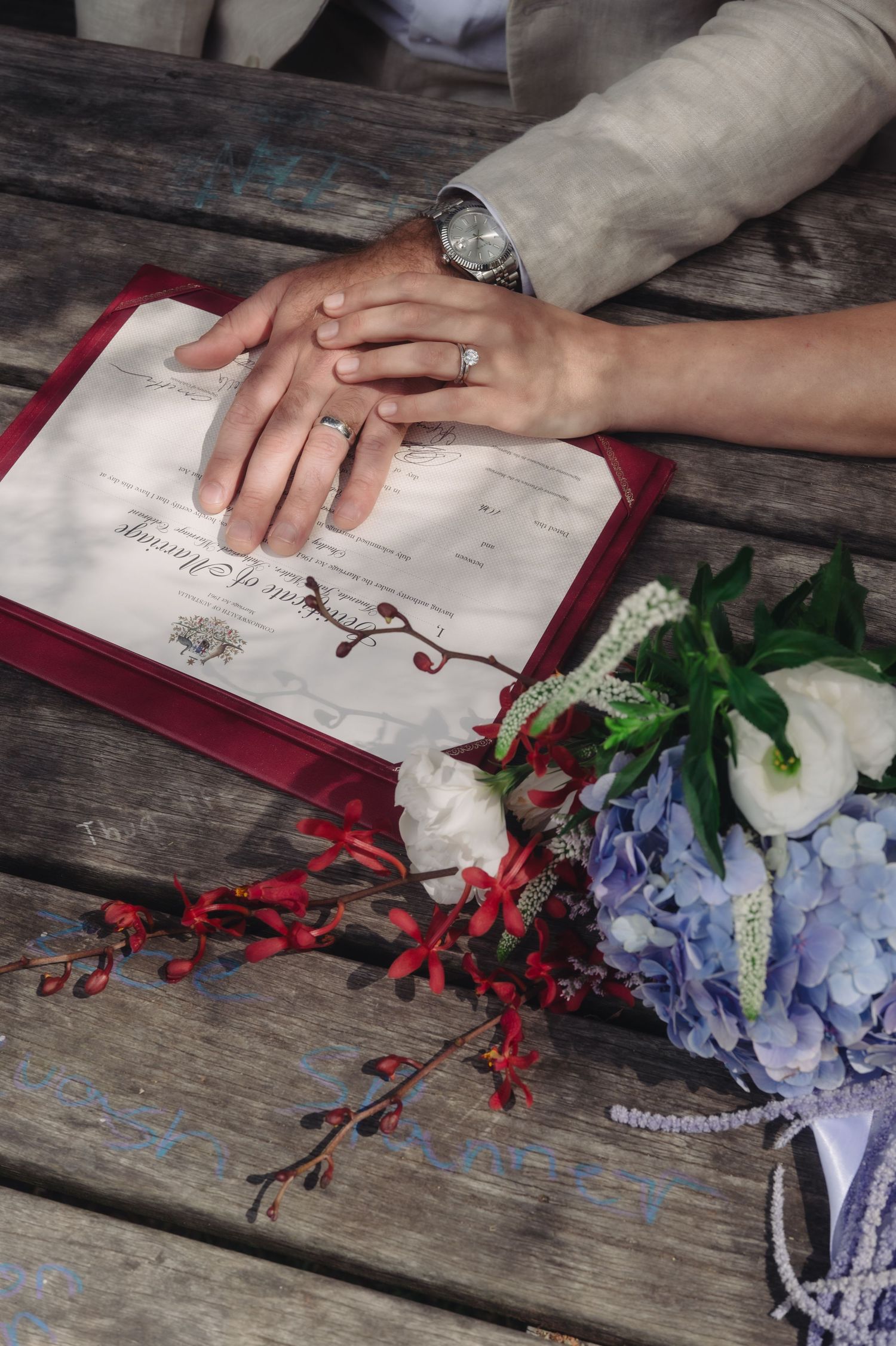 Newlyweds place hands on marriage certificate with wedding rings visible, beside a blue hydrangea bouquet on rustic wood.