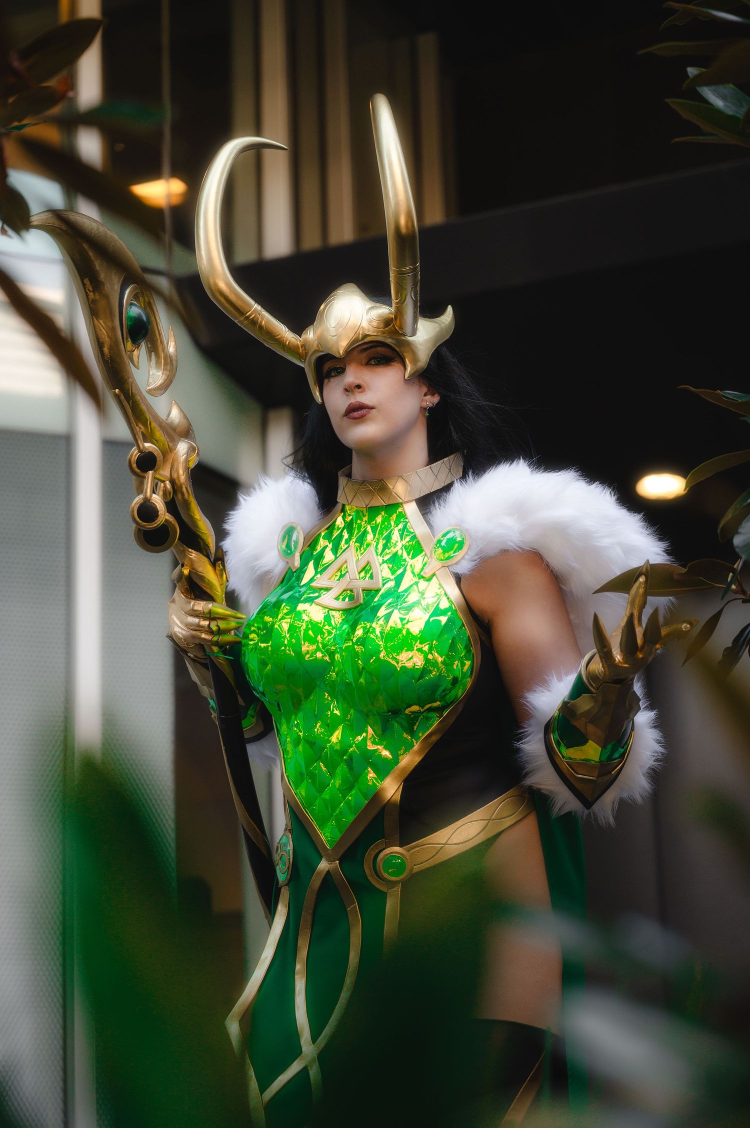 Woman cosplaying as Loki in green holographic armor, gold horned helmet, fur collar, holding golden staff at convention.