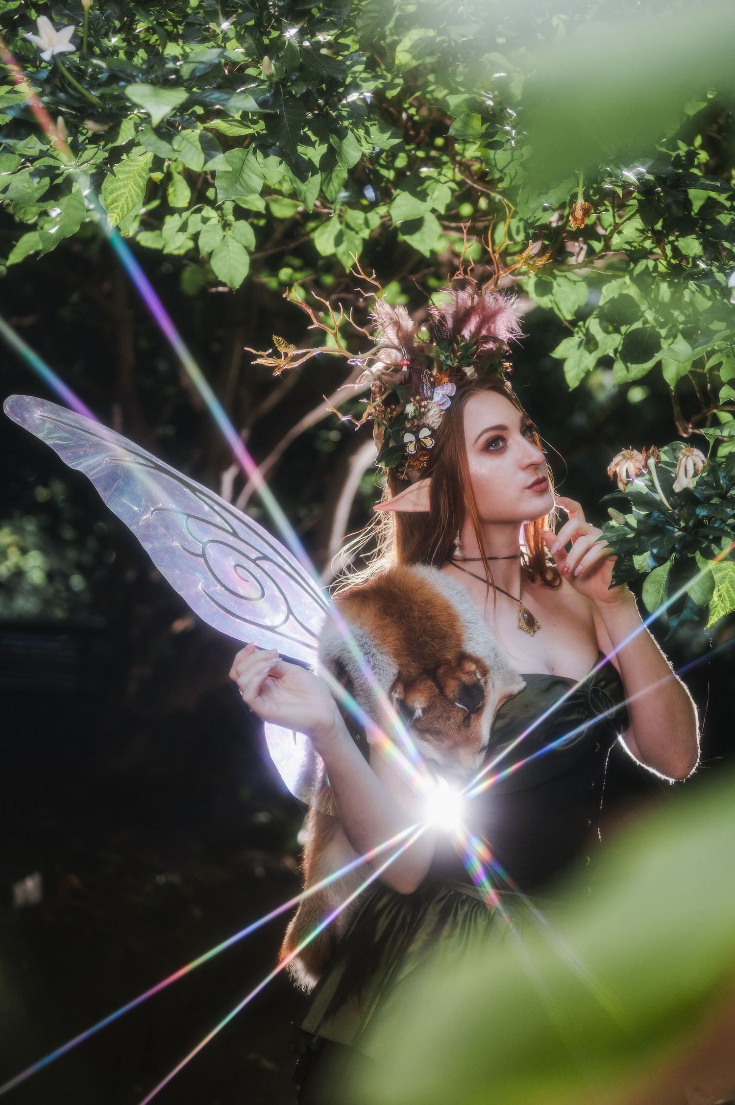 A fairy cosplay woman with wings and floral crown holds a small animal in an enchanted forest with rainbow light rays.