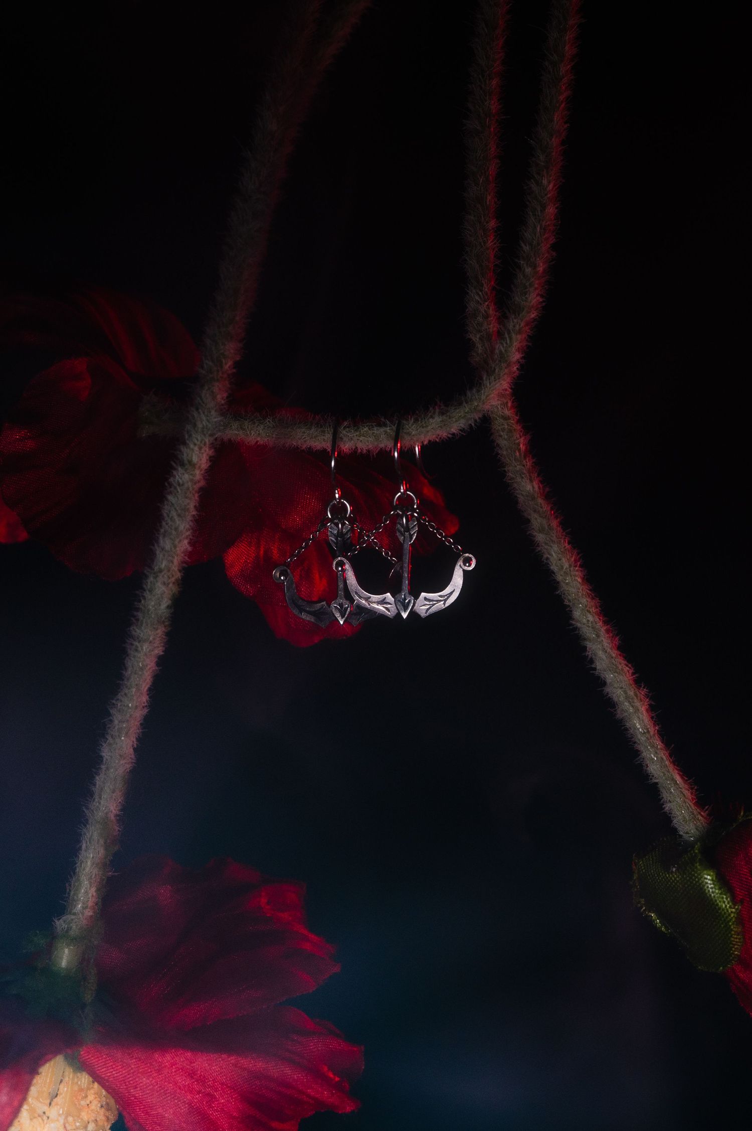 Silver anchor charm pendant hanging from red rope cord, surrounded by dark red flowers on black background.