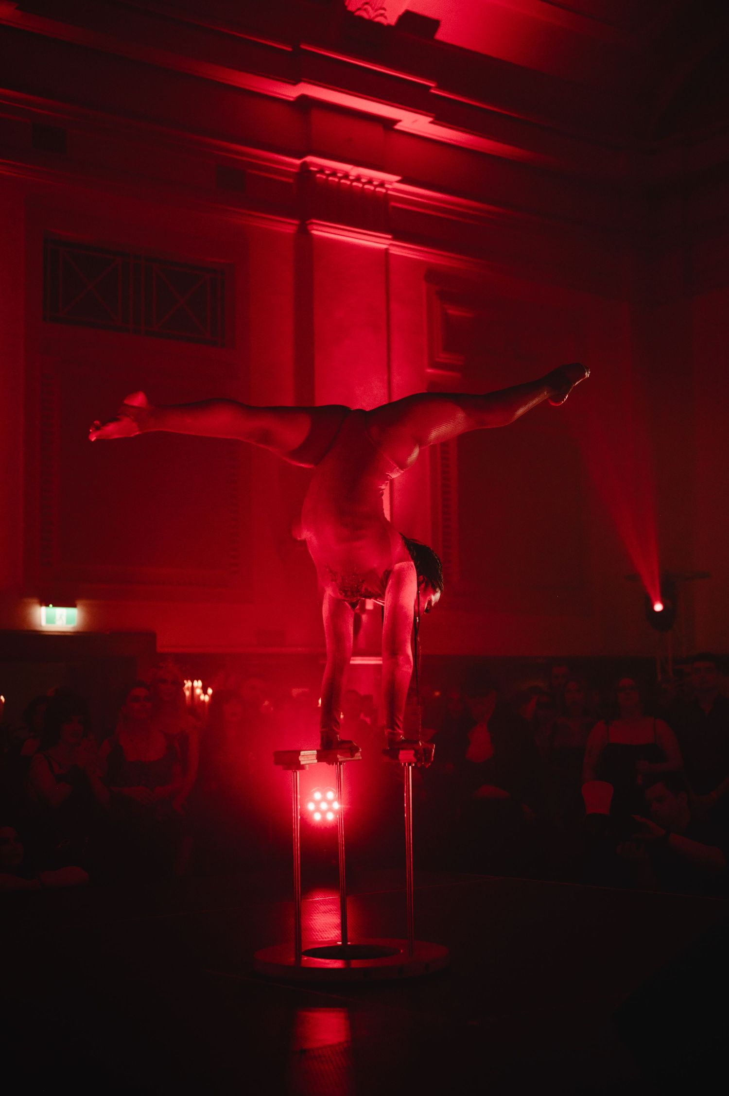 Acrobat performing a handstand split on a small pedestal in a red-lit hall while a seated audience watches.