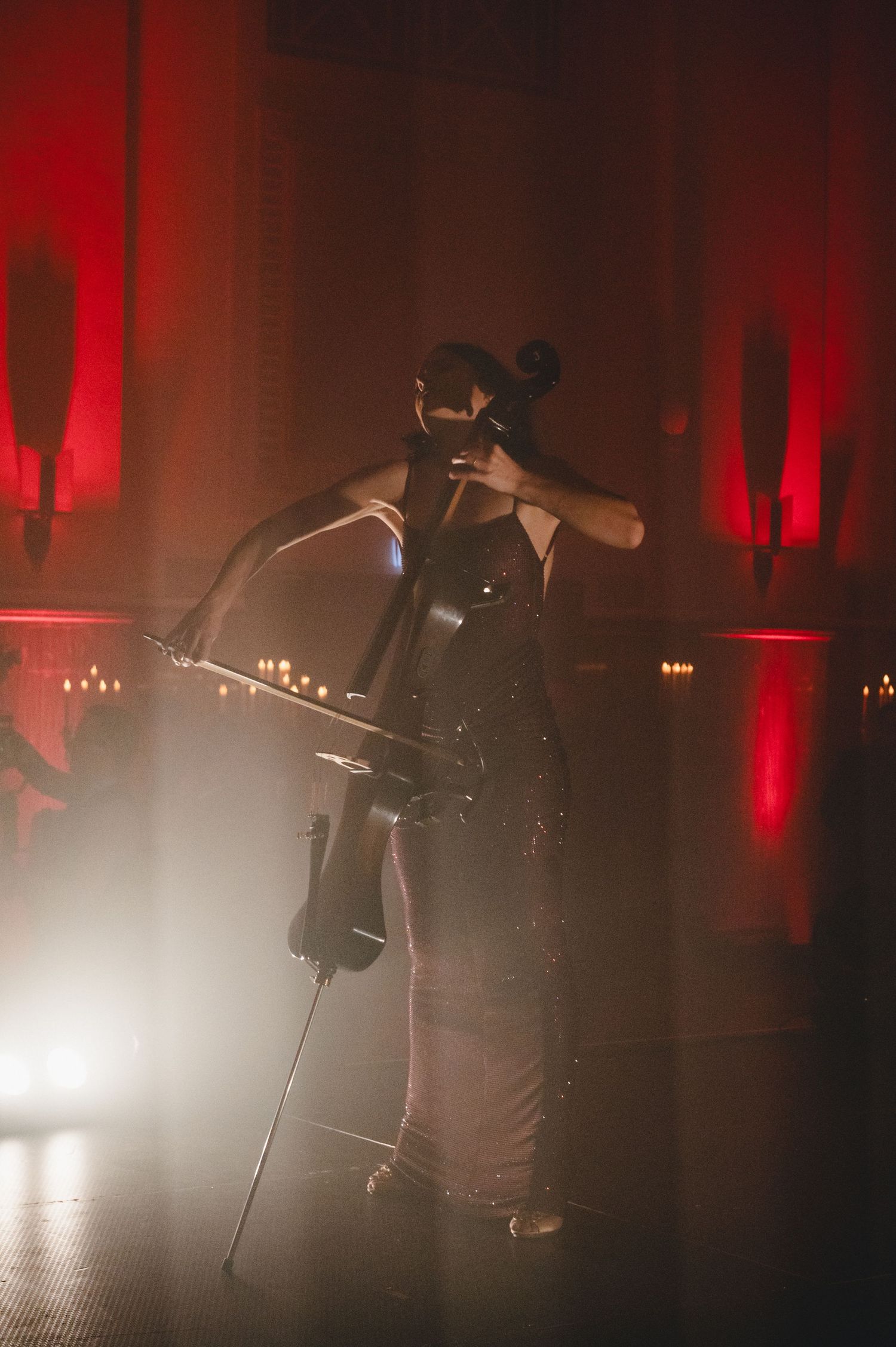 Cellist performing on stage in dramatic red lighting, wearing a mask and elegant gown in a moody atmospheric venue.