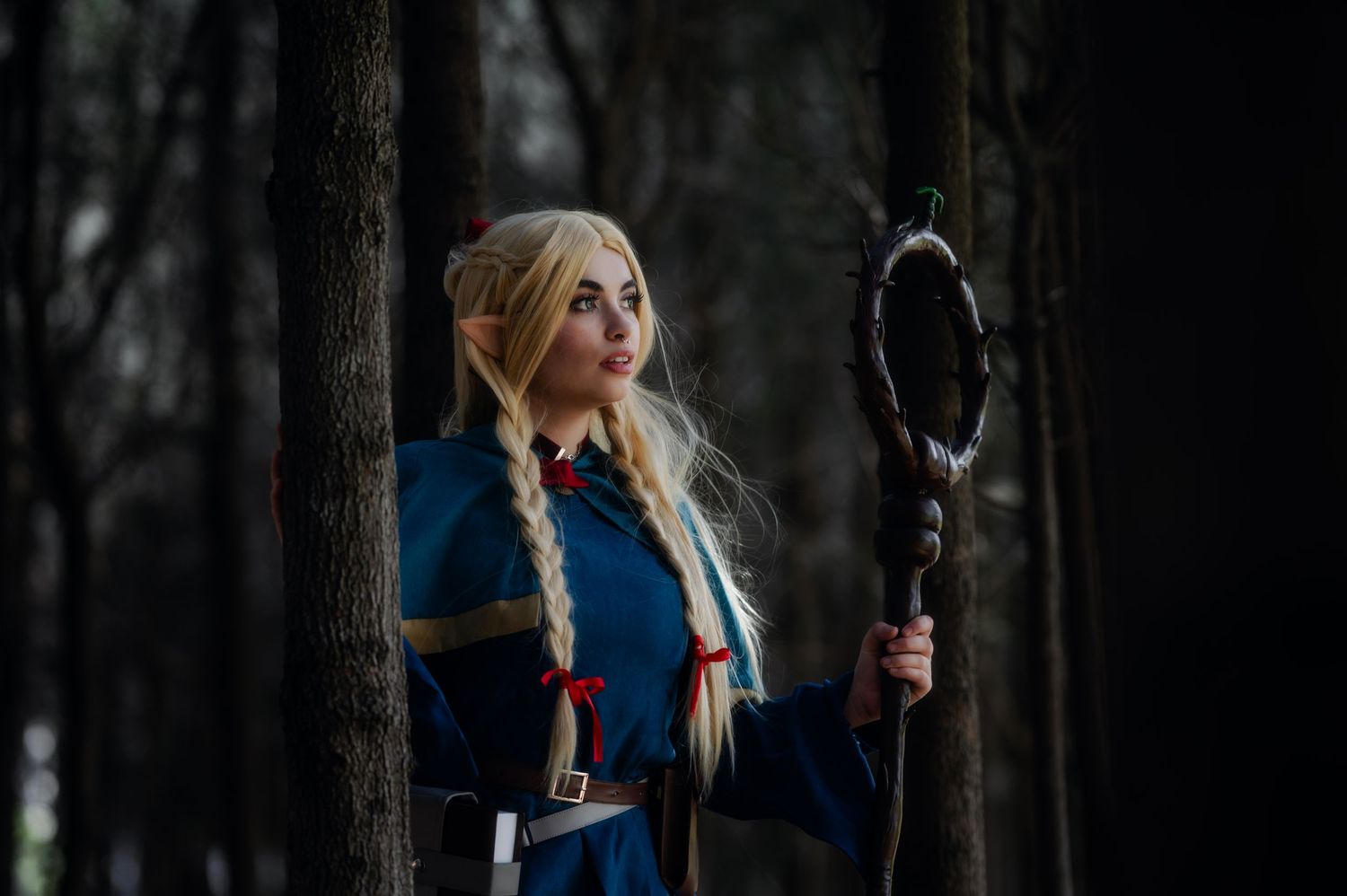 Fantasy cosplay character in dark forest wearing blue outfit and holding wooden staff.