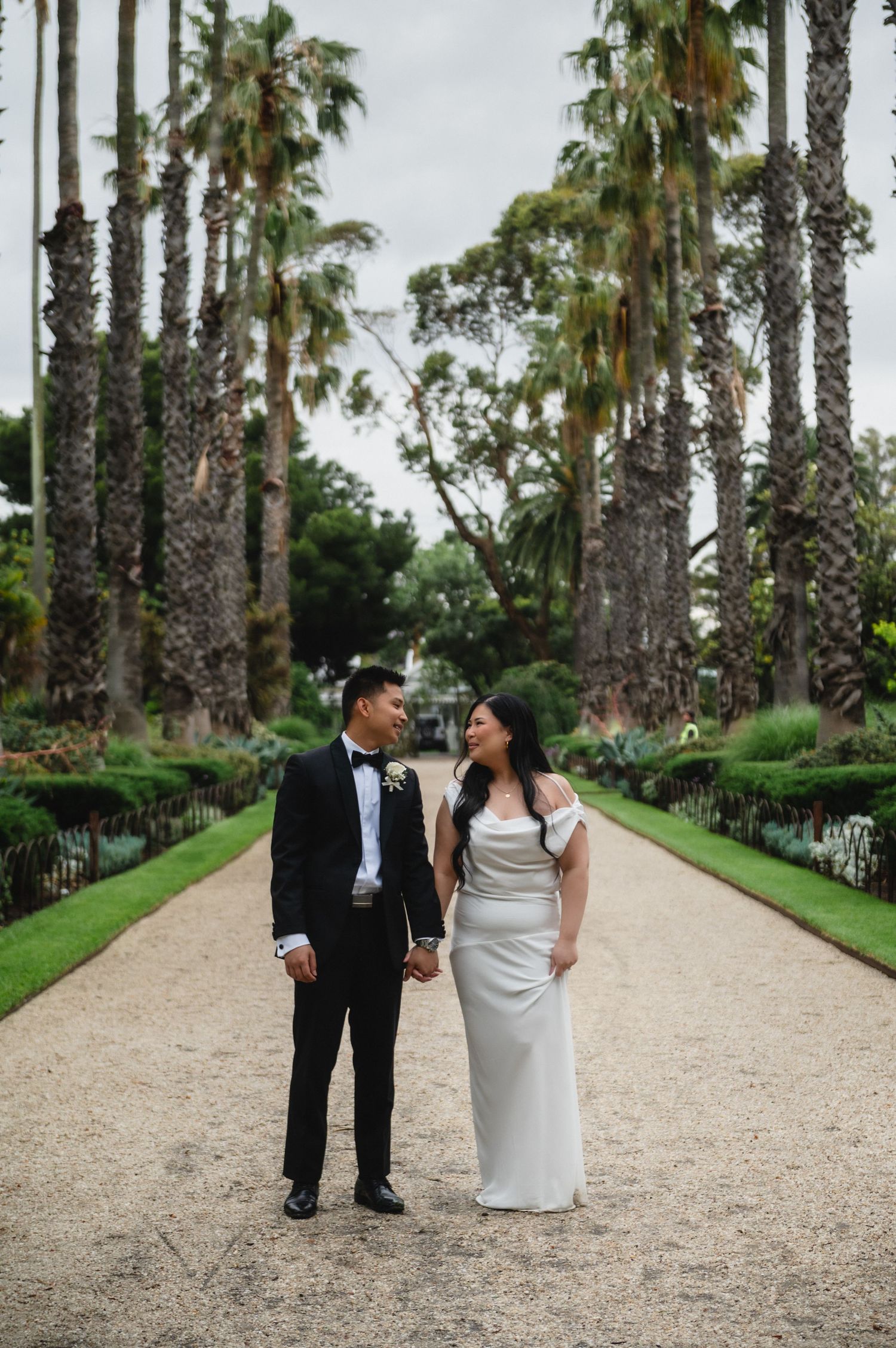 A couple in wedding attire walks together on a palm tree-lined path at an outdoor venue.