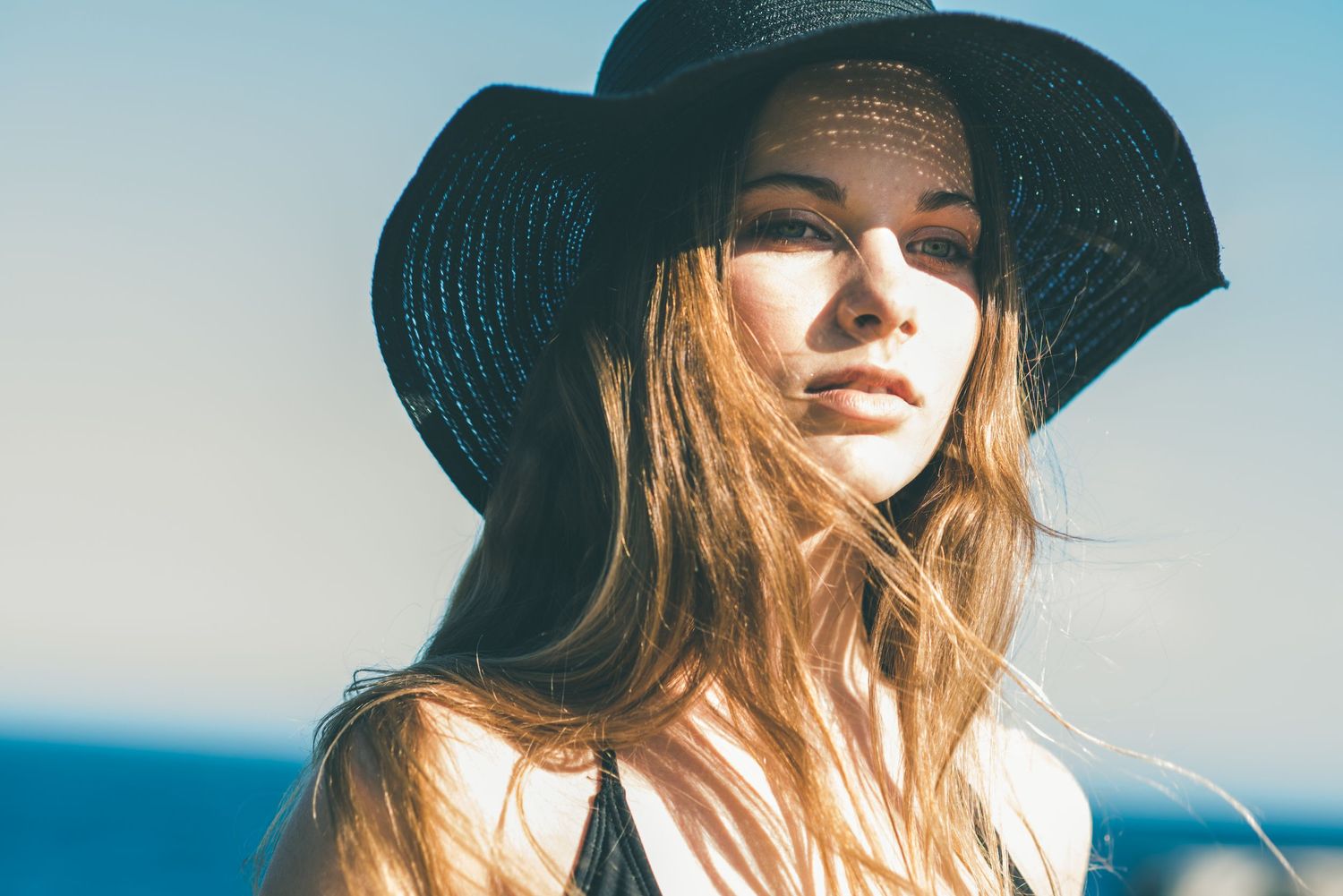 Young woman with long hair wearing a wide-brim black hat outdoors on a sunny day near the ocean.