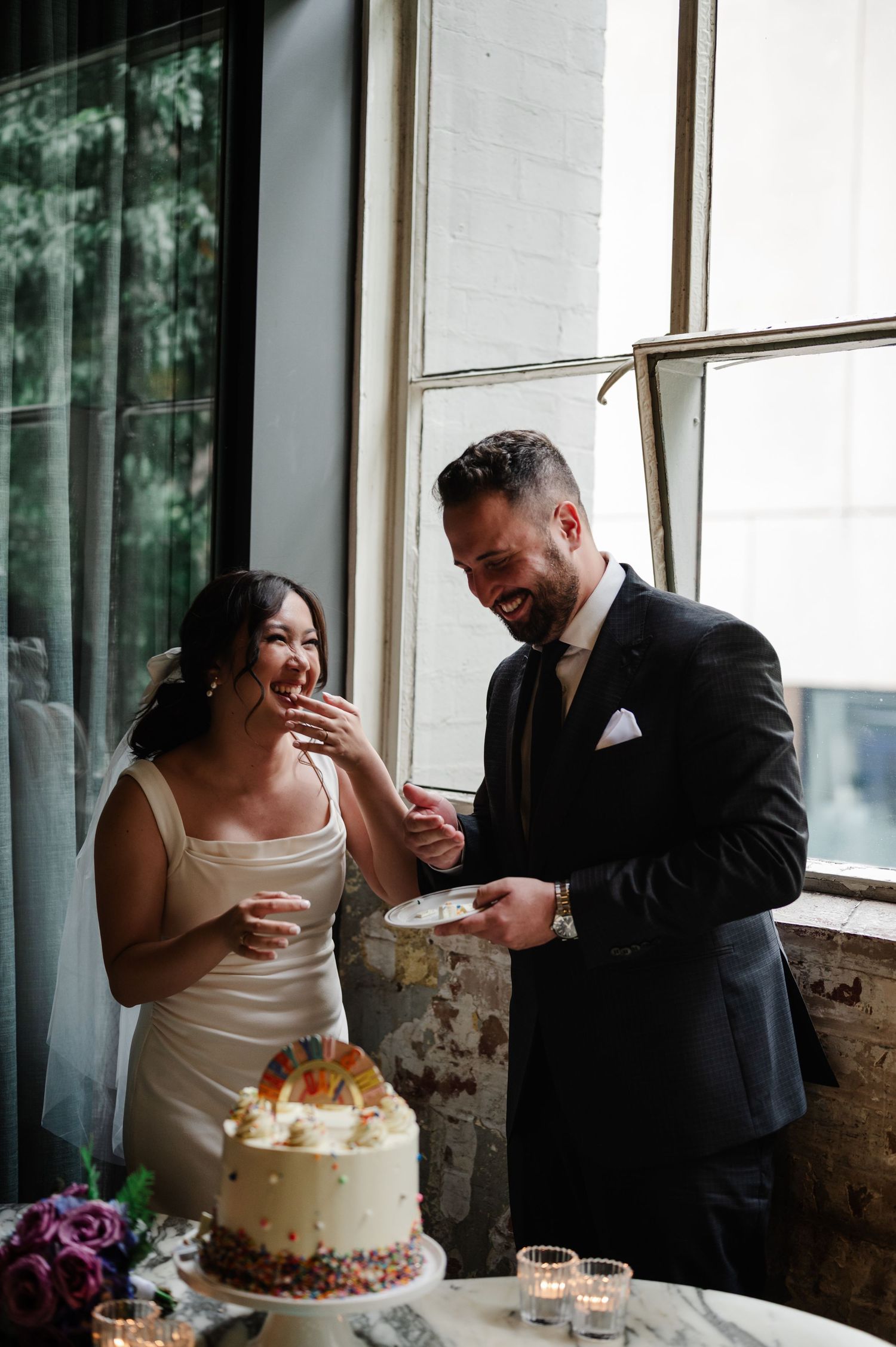 Couple sharing cake at their wedding reception by large windows.