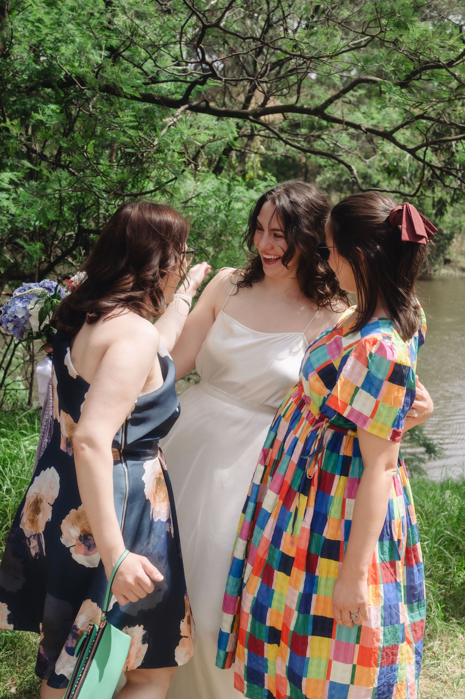 Bride in white dress laughing with two friends in floral and colorful patchwork dresses outdoors near trees.