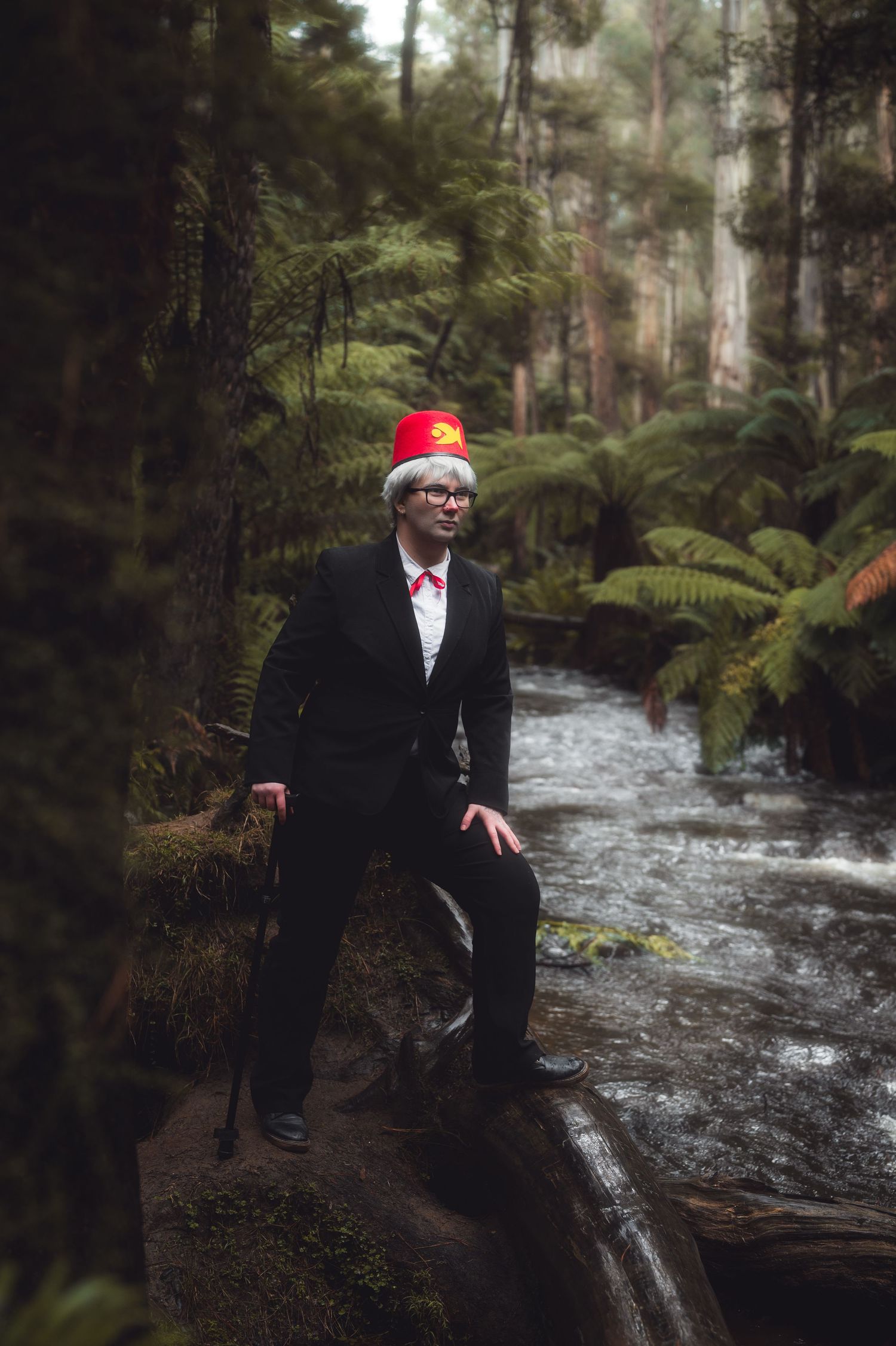 A figure in formal attire stands near a rushing stream in a lush forest landscape.