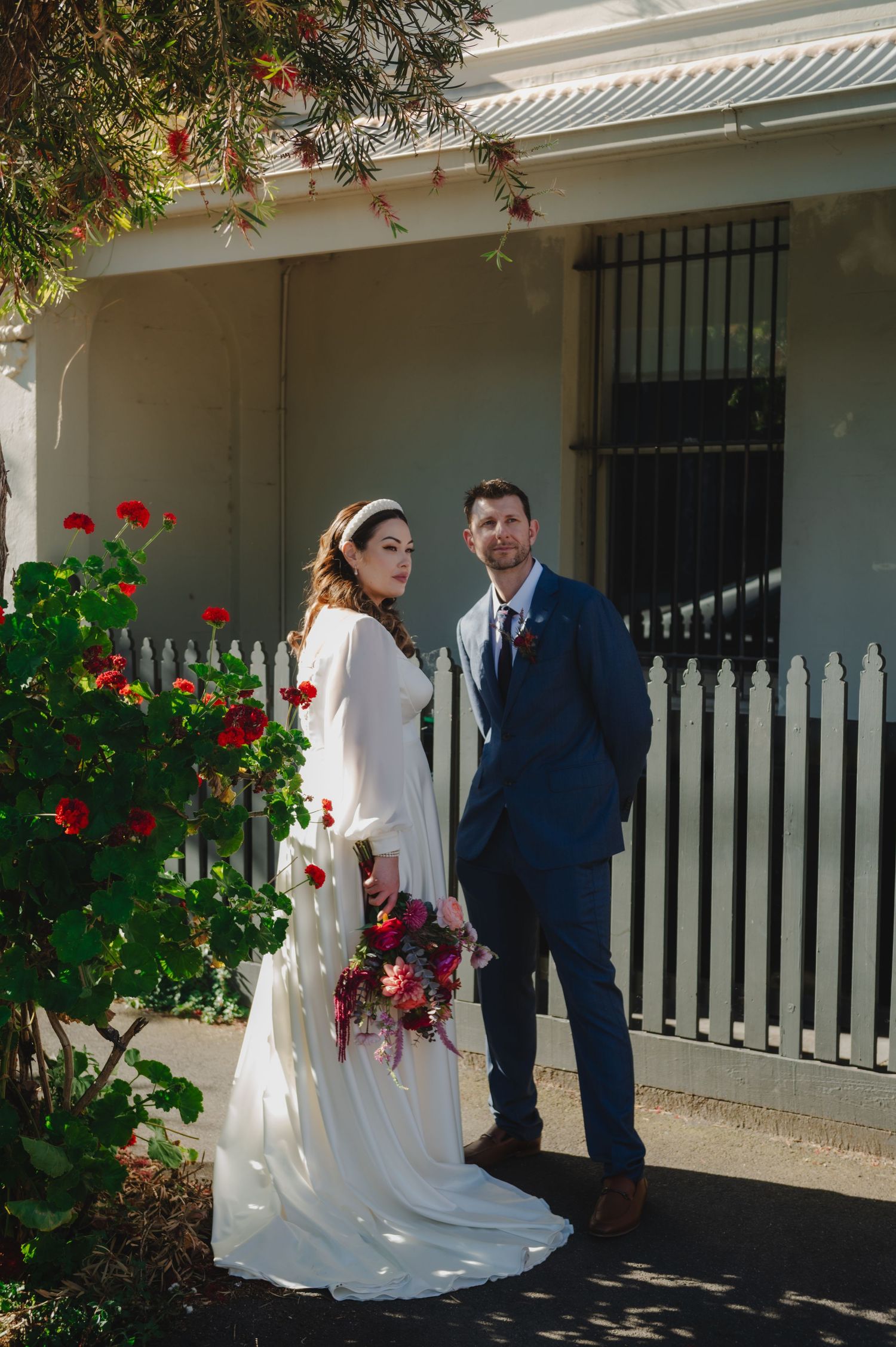 A wedding couple poses together by a white picket fence and blooming red flowers on a sunny day.
