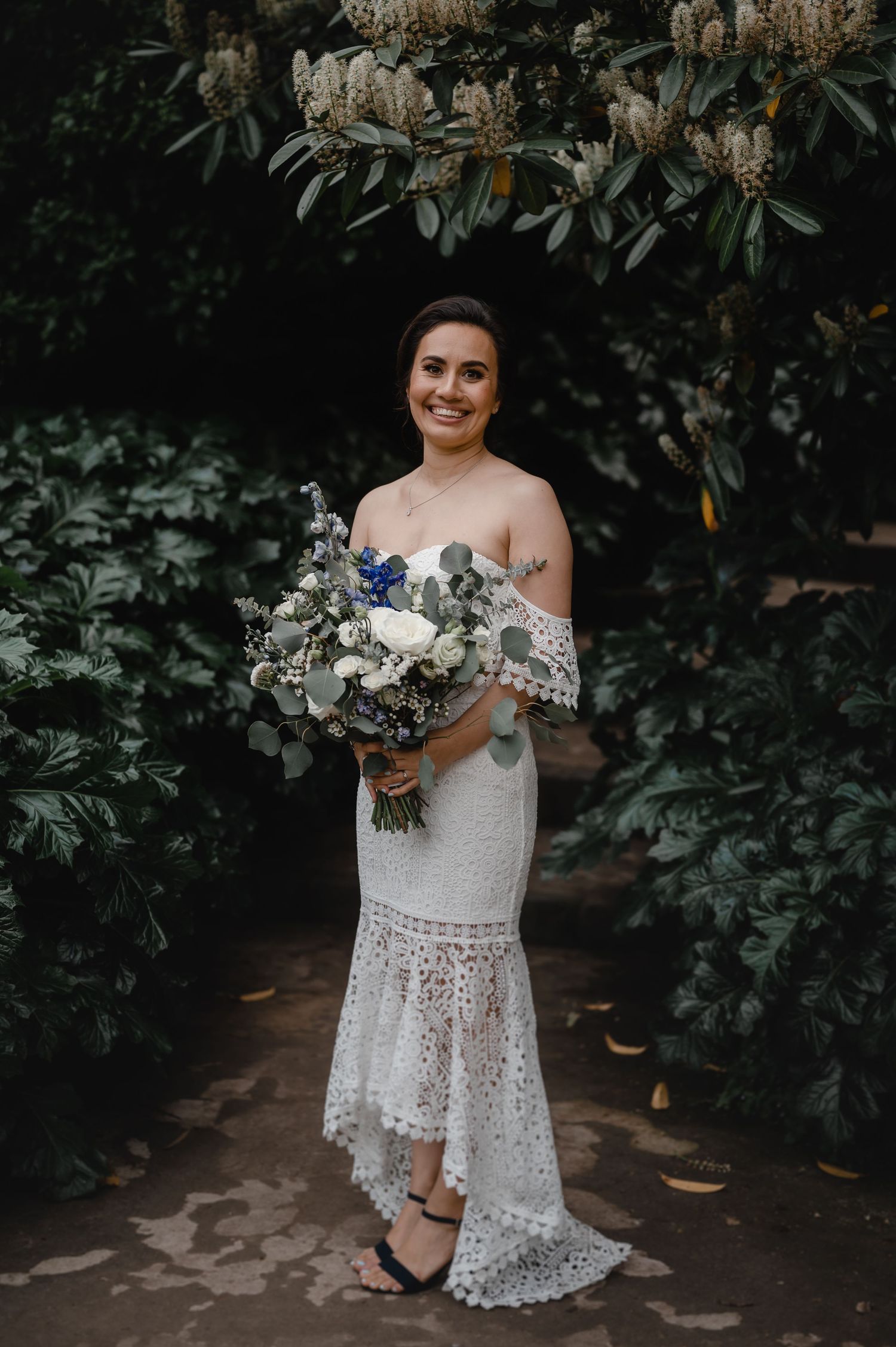 Elegant bridal portrait with lace dress and blue-white bouquet in garden setting.