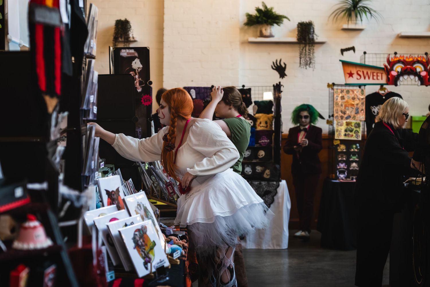 People in Halloween costumes browse vendor booths at an indoor convention or market event.