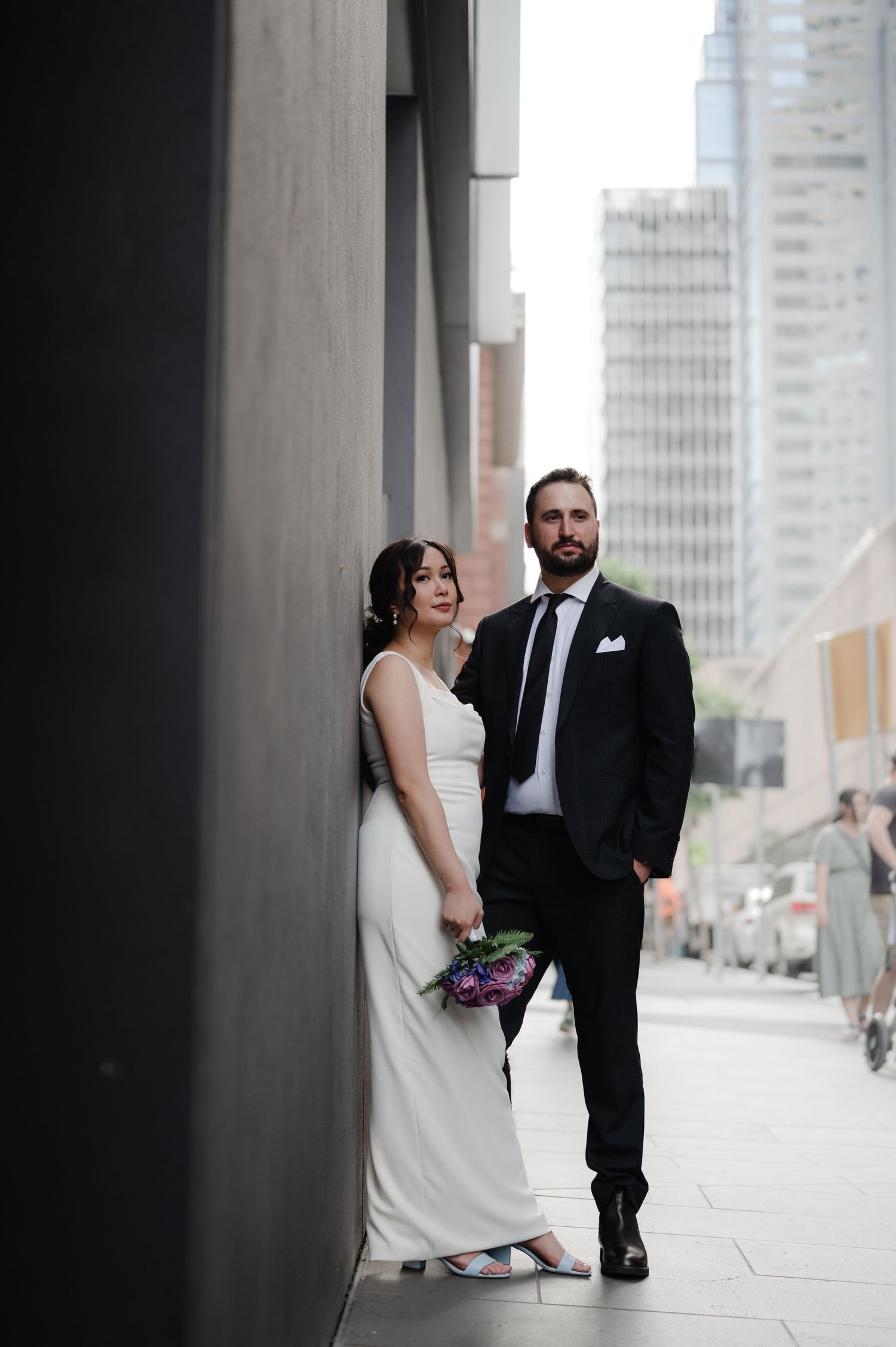 Modern wedding couple poses between urban columns with city skyline in background.