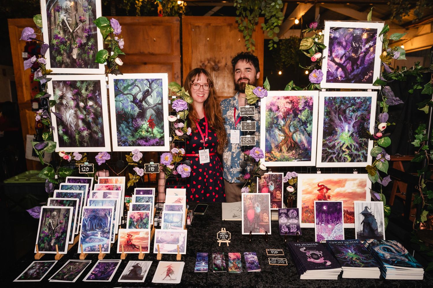 Collection of framed artwork and prints displayed on easels at an outdoor art market under string lights.
