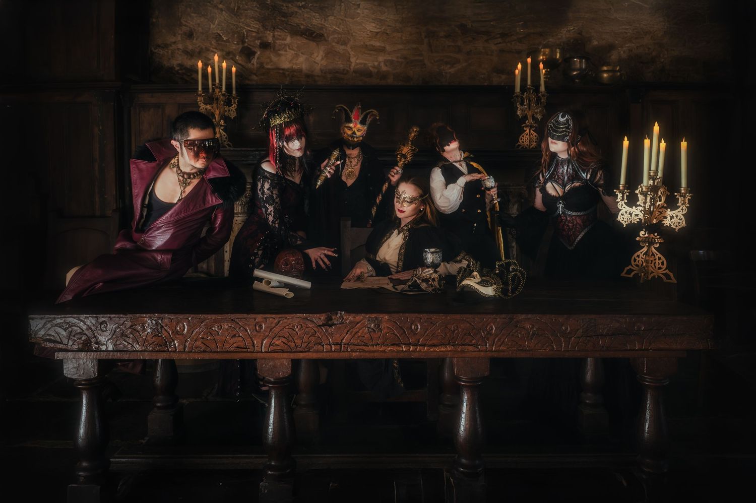 A group in Victorian gothic attire poses at a long table with candelabras in a dark chamber.
