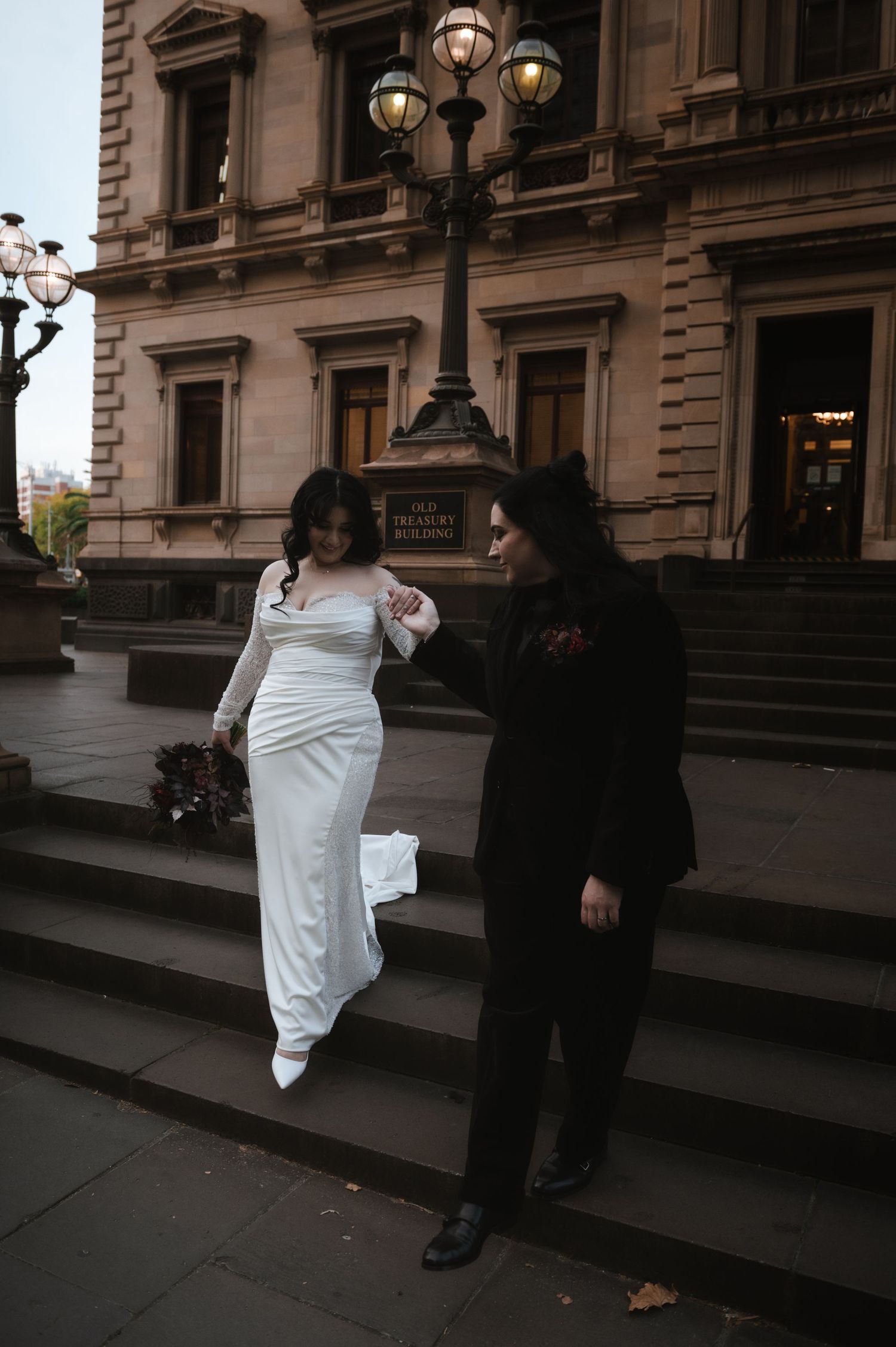 Wedding couple posing on stone steps outside a historic building with ornate lamp posts.