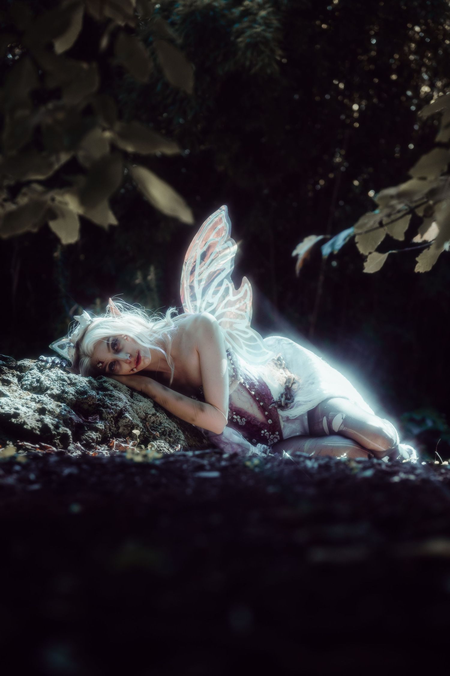 A glowing fairy with white hair and pink wings rests on mossy rocks in an enchanted forest setting.