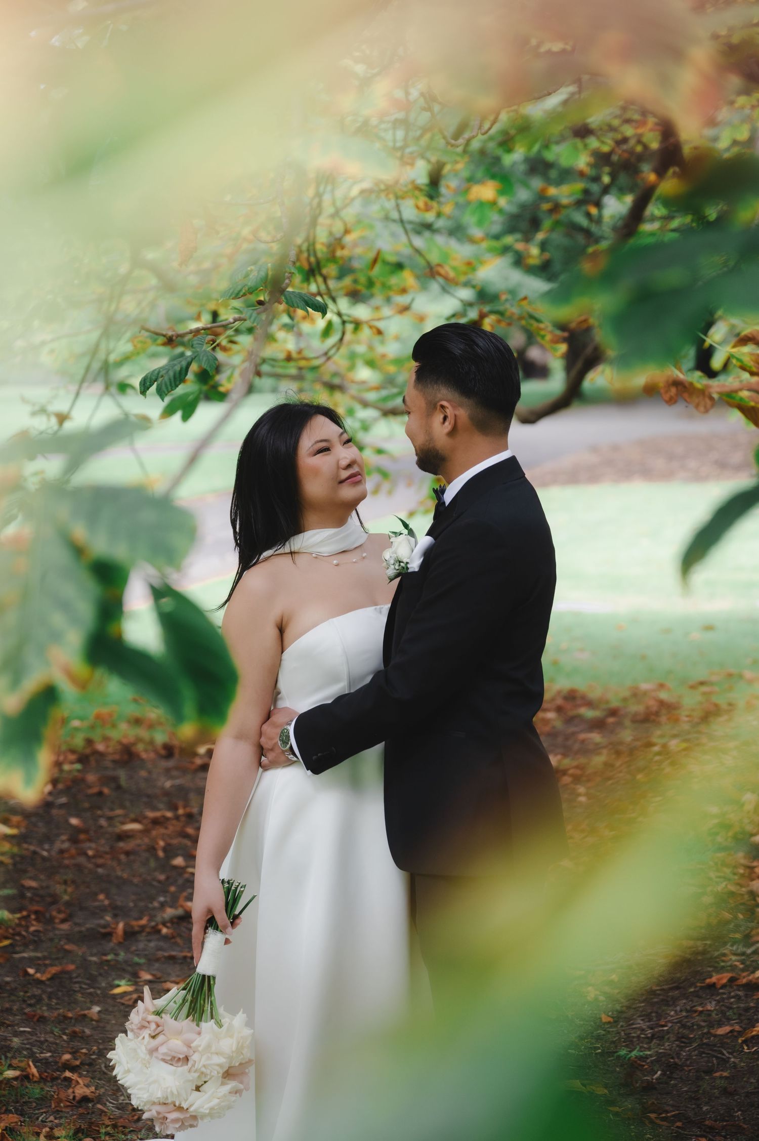 Bride in white strapless gown holding bouquet stands with groom in black tuxedo under lush green trees.