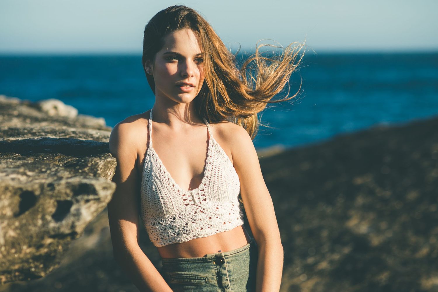Young woman in white crochet crop top and jeans posing on rocky coastal shore with ocean in background.