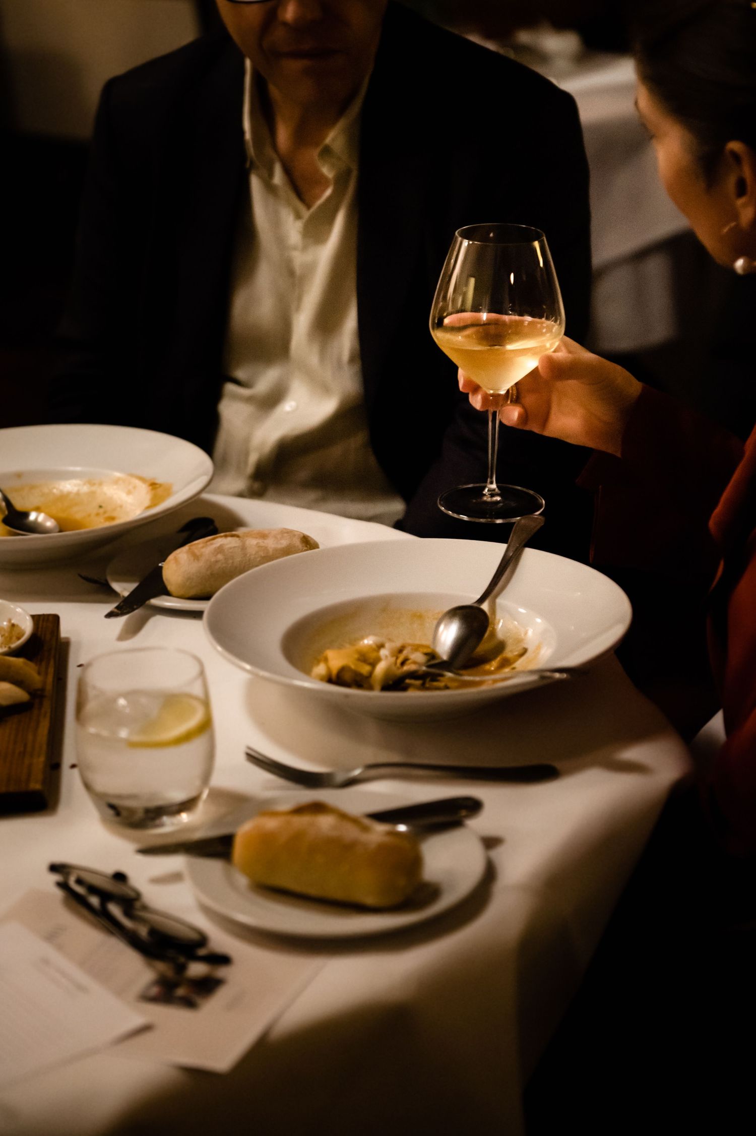 Diners enjoy soup and white wine at an elegant restaurant dinner table with bread.