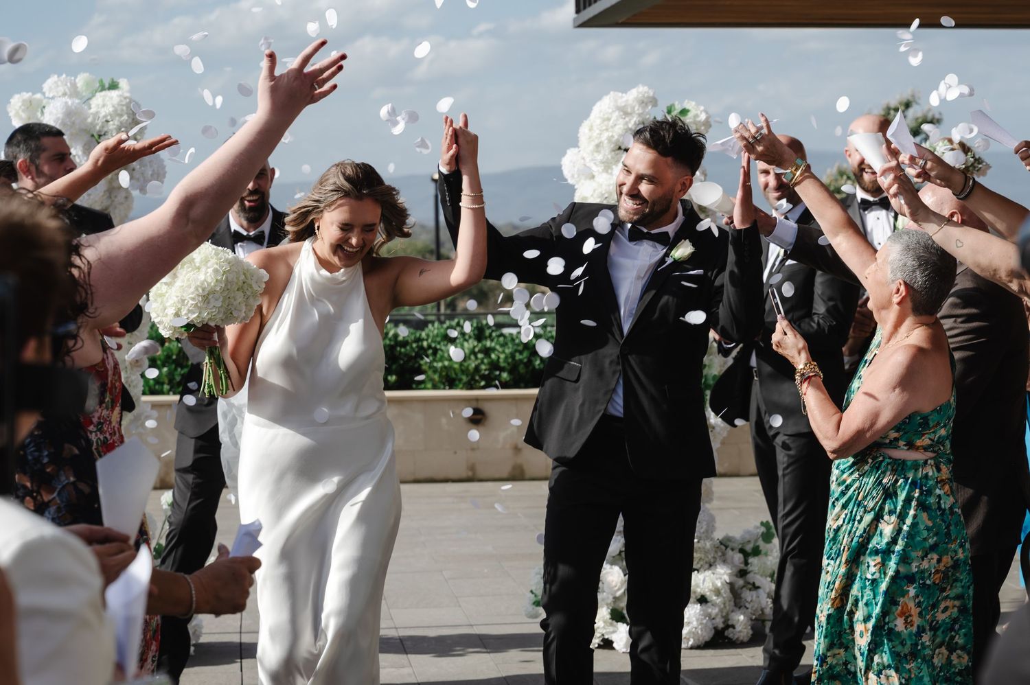 Wedding celebration with guests throwing confetti outside on a sunny day.