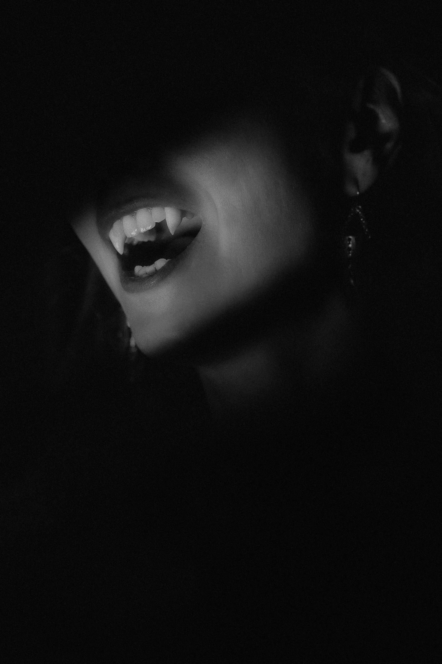Black and white portrait of a woman with vampire fangs and dangling earrings emerging from dramatic shadows.