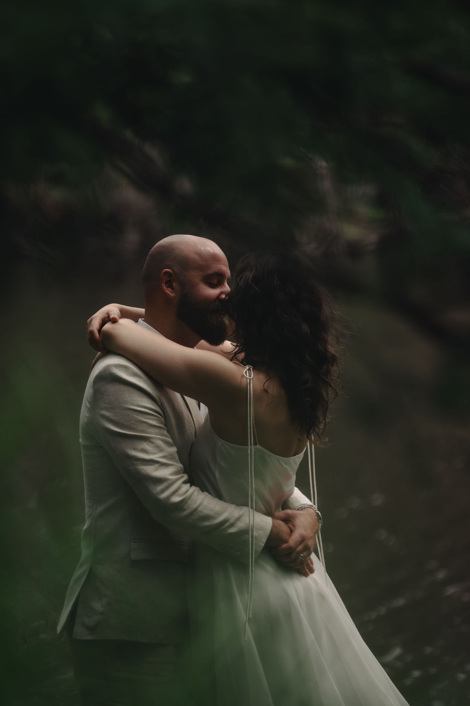 A couple shares a romantic embrace in a lush forest setting, the bride in a white gown and groom in a light suit.