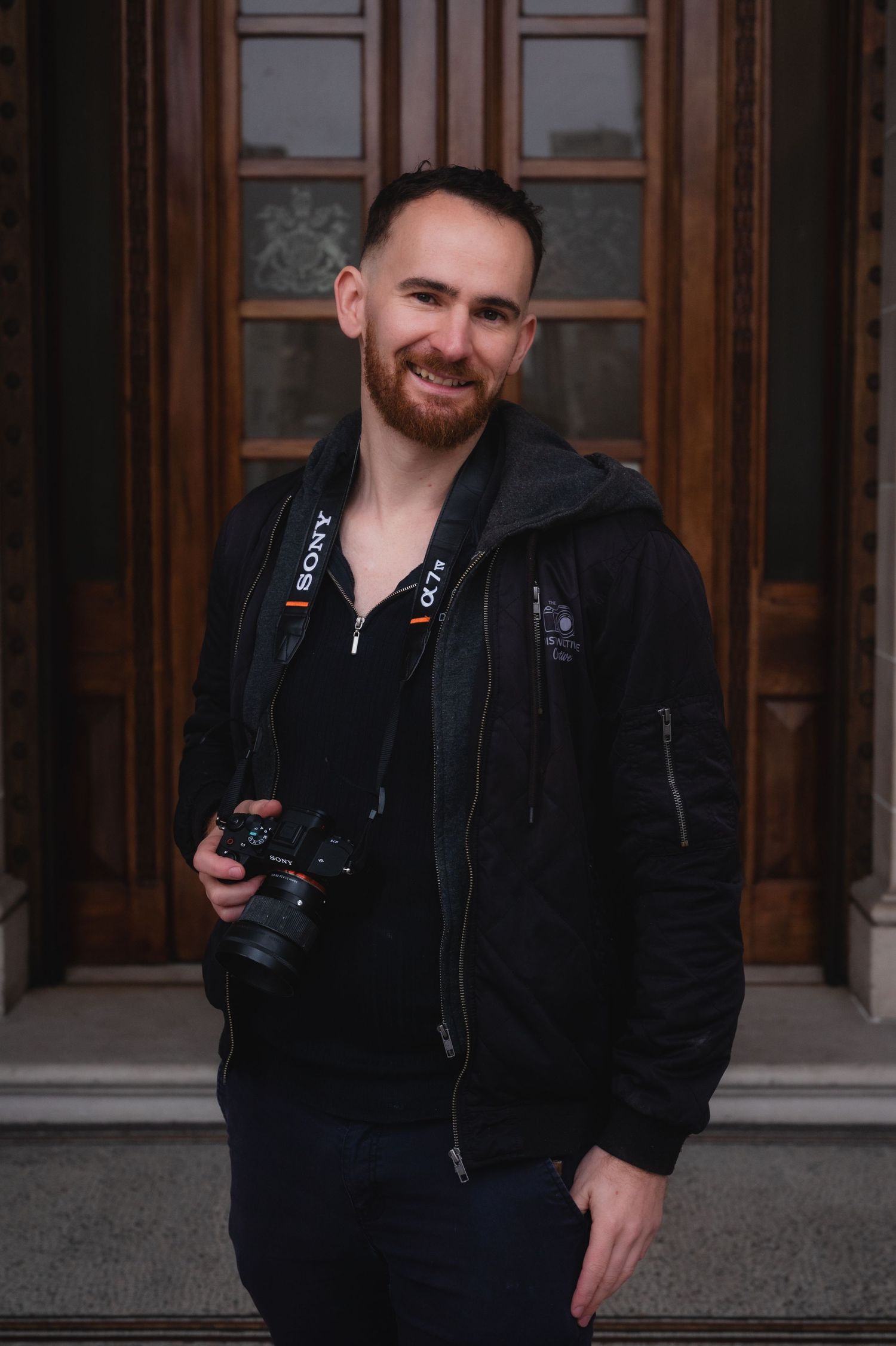 A smiling man with a beard holds a Sony camera while wearing a black hoodie, standing in front of ornate wooden doors.