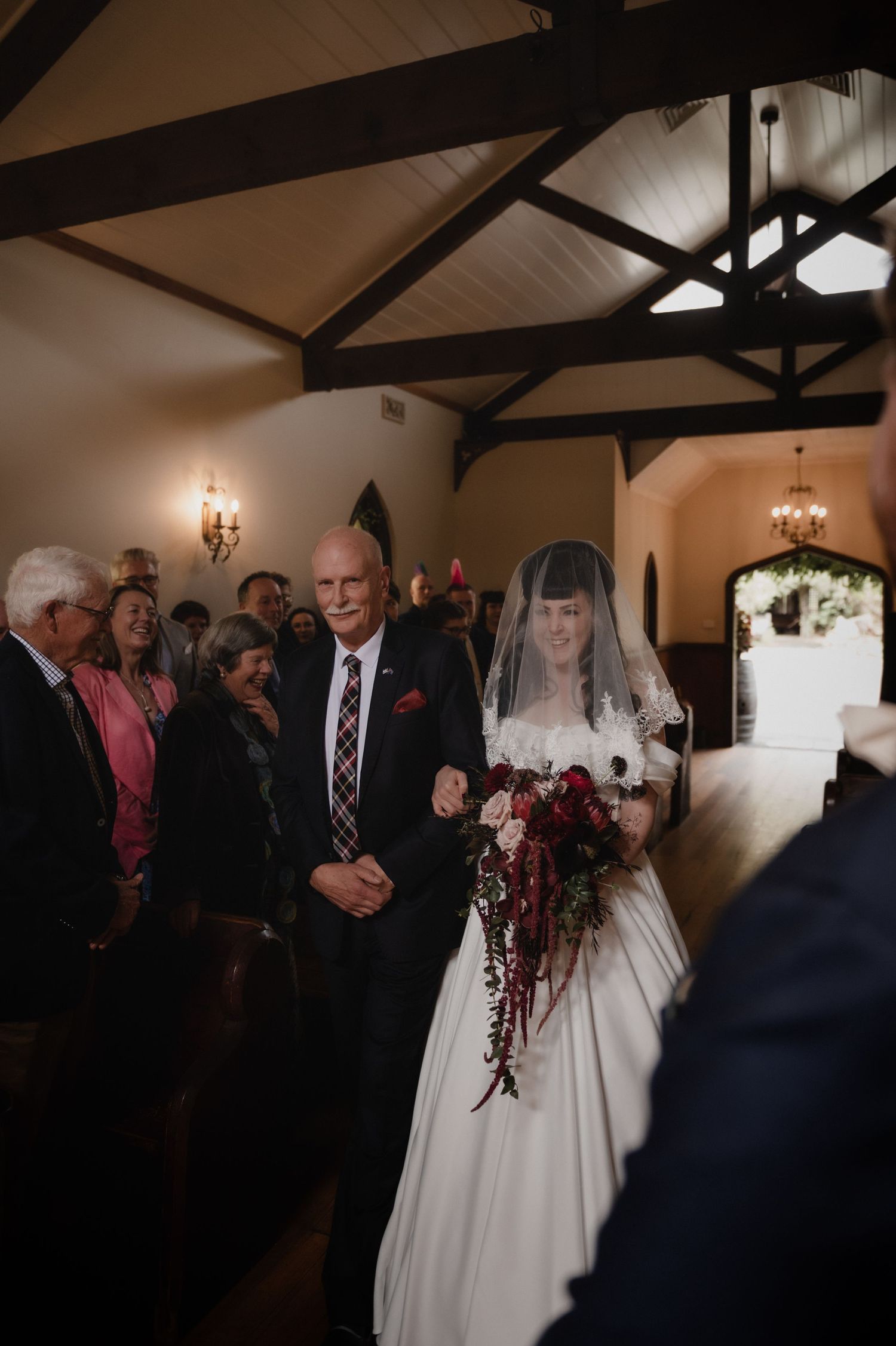Bride walking down the aisle with her father in a rustic chapel, wearing a white gown and veil, holding a burgundy bouquet.