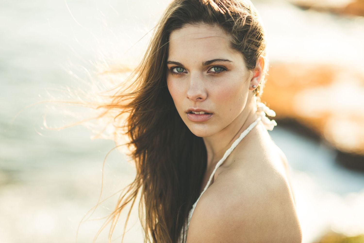 Young woman with long brown hair poses outdoors near rocky coast, wind-blown hair, natural makeup, soft golden light.