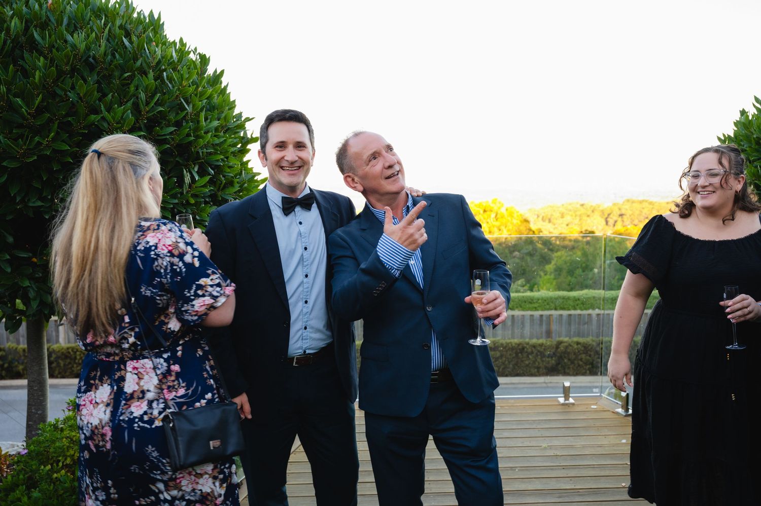 A group of formally dressed guests share a laugh together at an outdoor evening event on a deck.